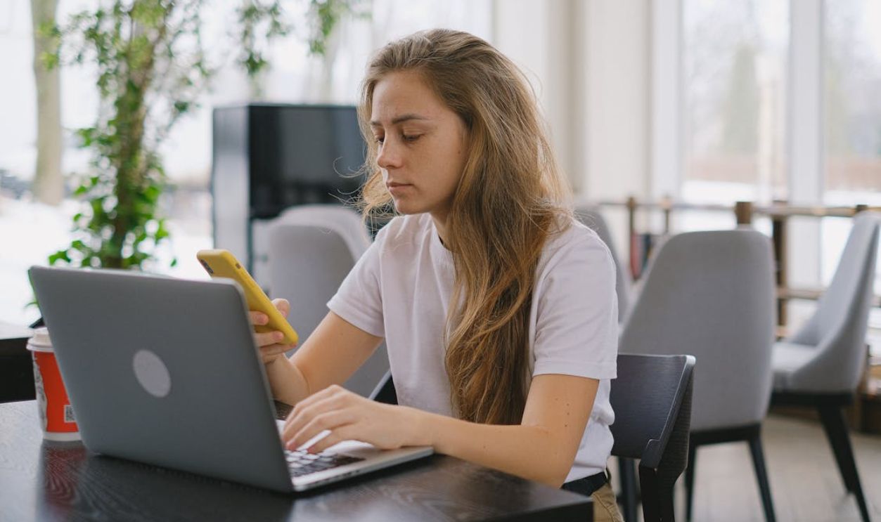 Focused woman using a laptop and smartphone at a cafe, showcasing modern multitasking.