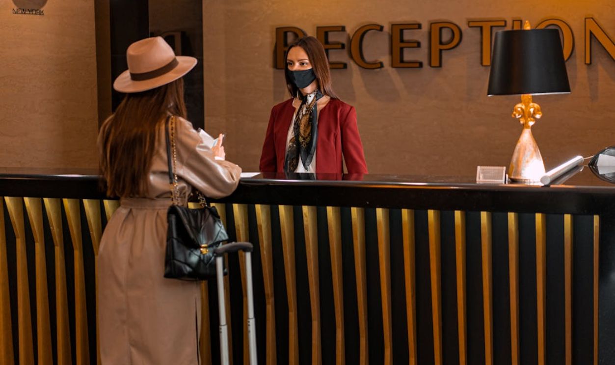 A woman speaks to a receptionist wearing a mask at a hotel reception.