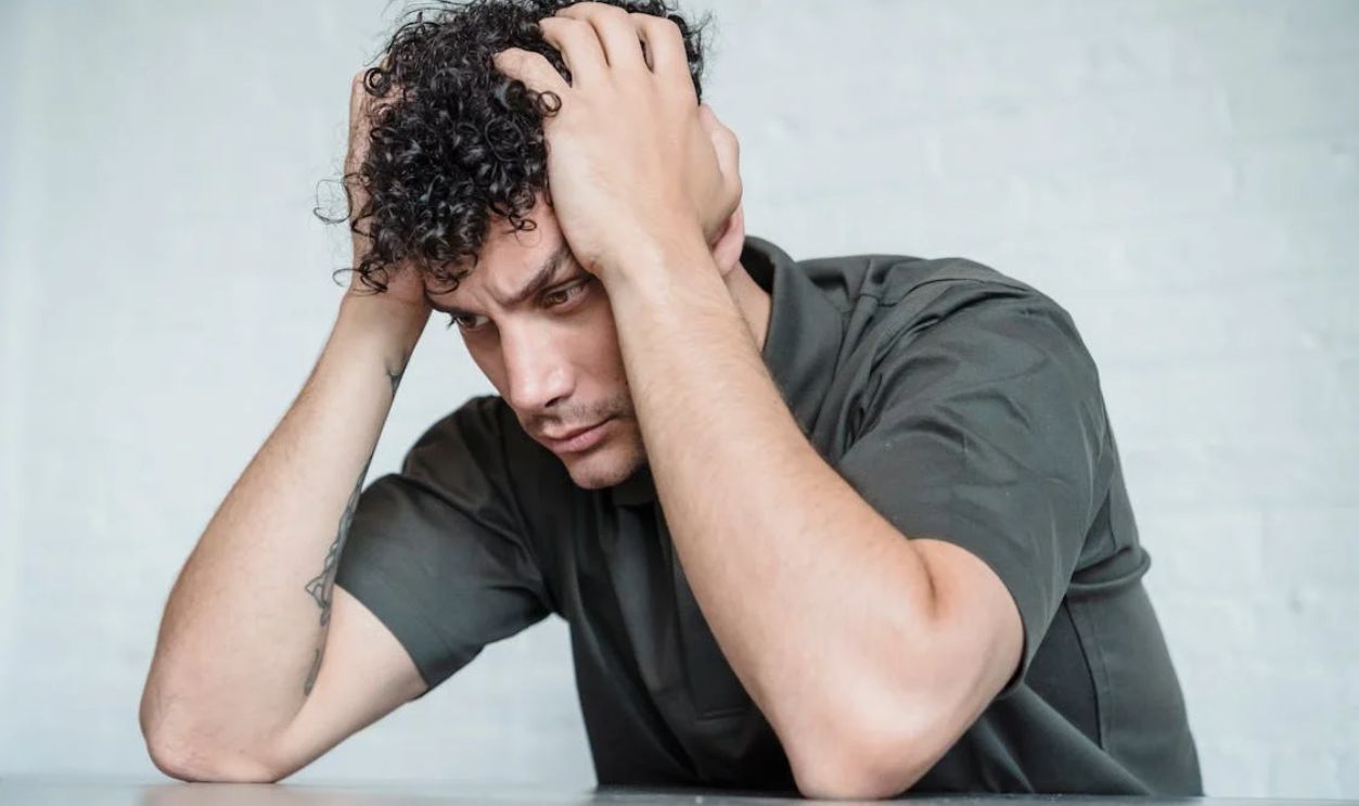 A young man with curly hair looks stressed as he sits with his hands on his head in contemplation.