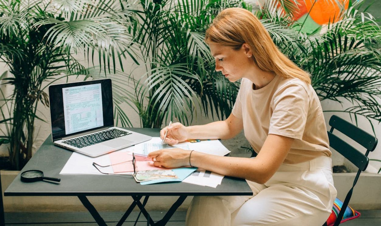 A woman deeply concentrated on work at her laptop amidst indoor greenery and documents.