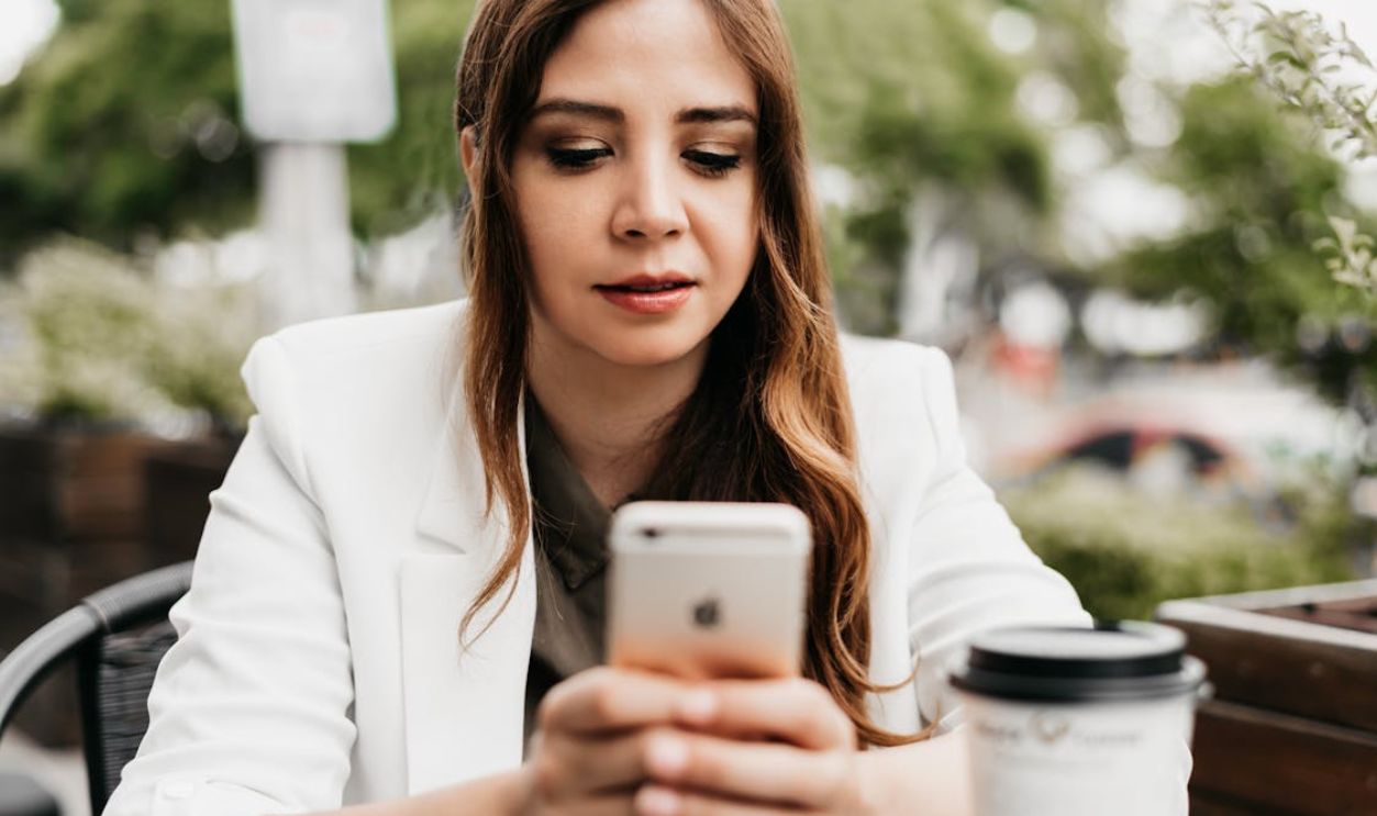 Woman in a white blazer using a smartphone at an outdoor cafe with coffee.