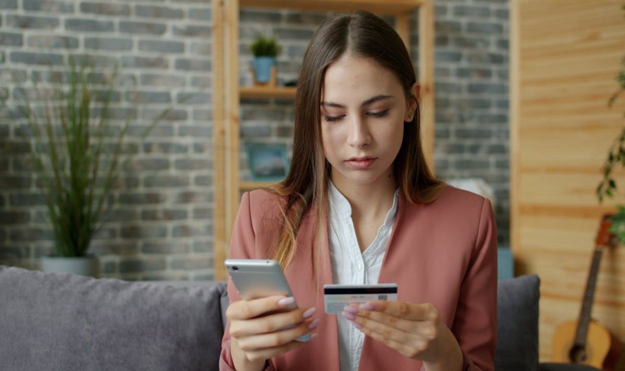 A young woman in a pink blazer shops online using a smartphone and credit card indoors.