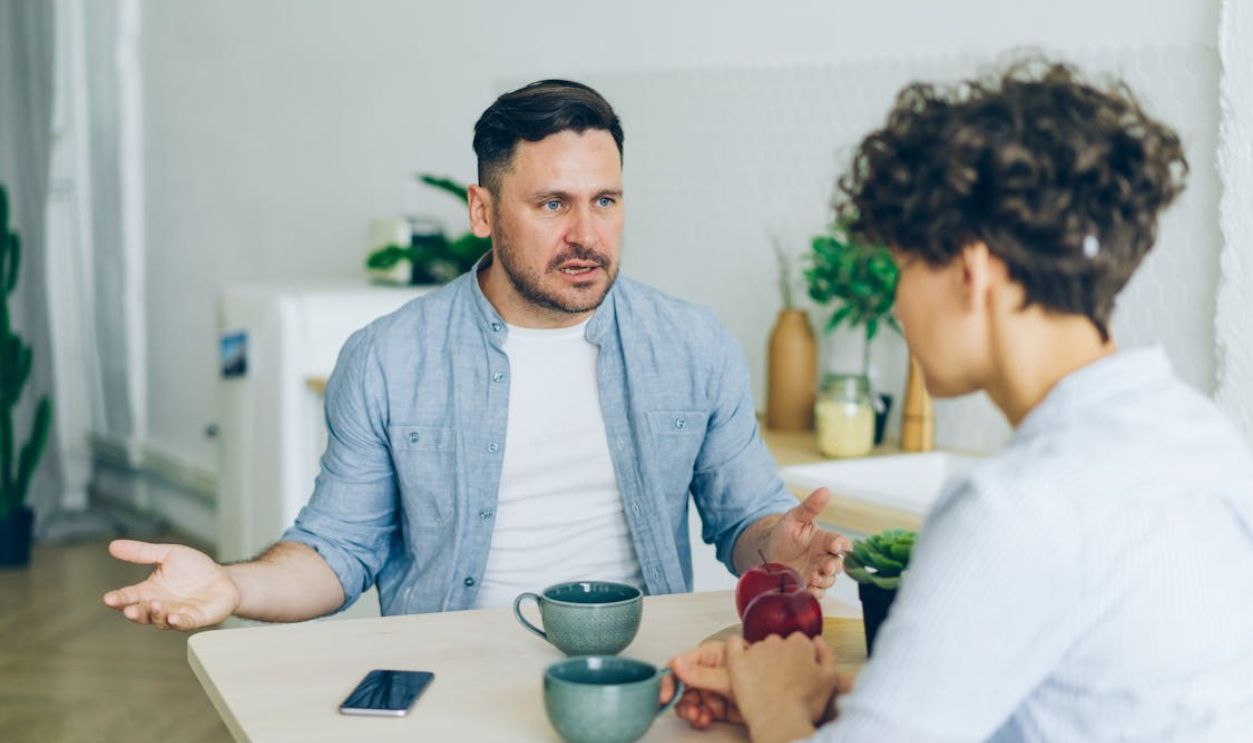 Two adults engage in a conversation at a kitchen table with coffee cups.