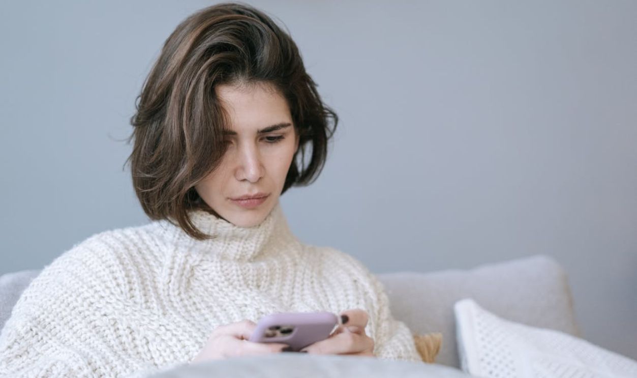 Woman in cozy knit sweater using a smartphone indoors, expressing a thoughtful mood.
