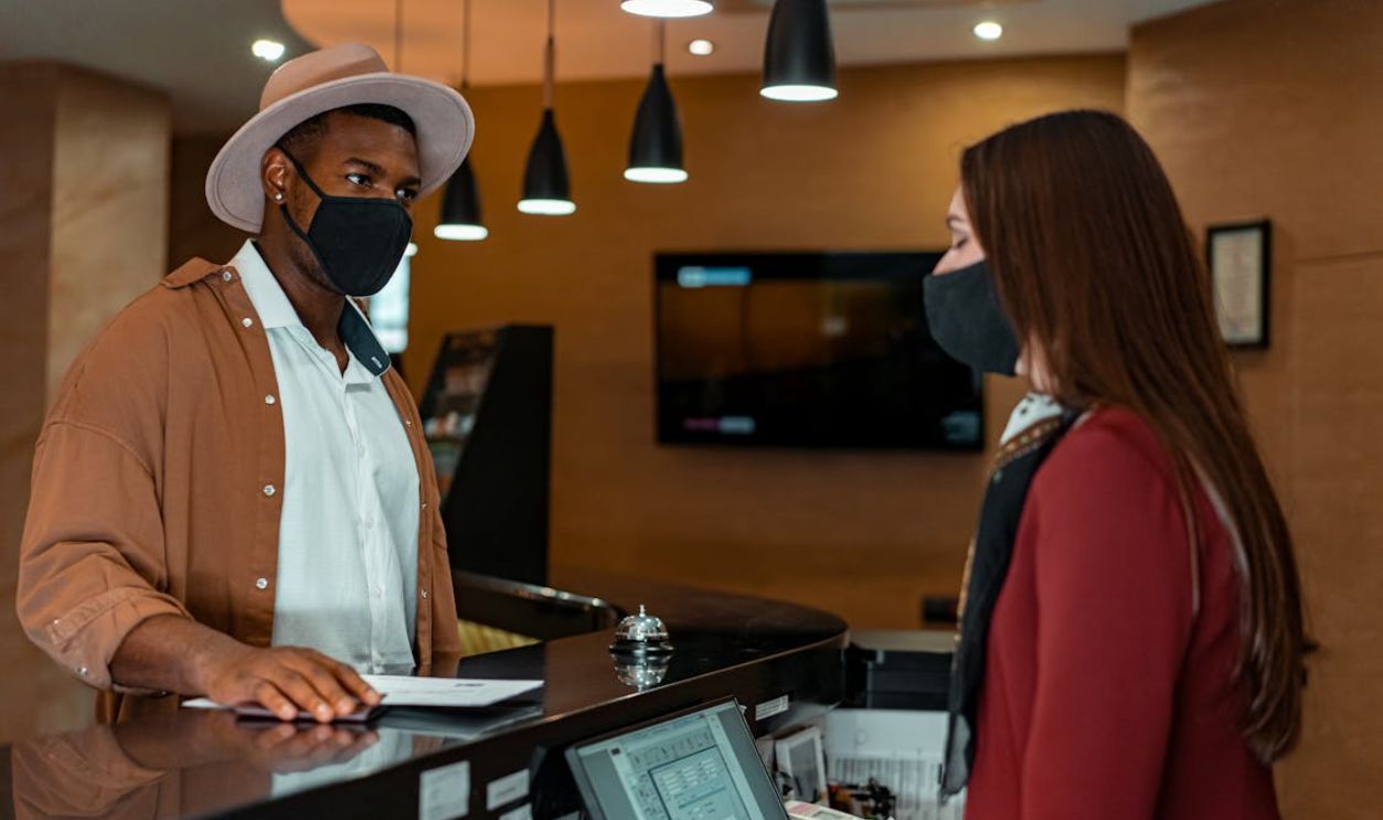 A man and woman in face masks checking in at a hotel reception, emphasizing safety protocols.