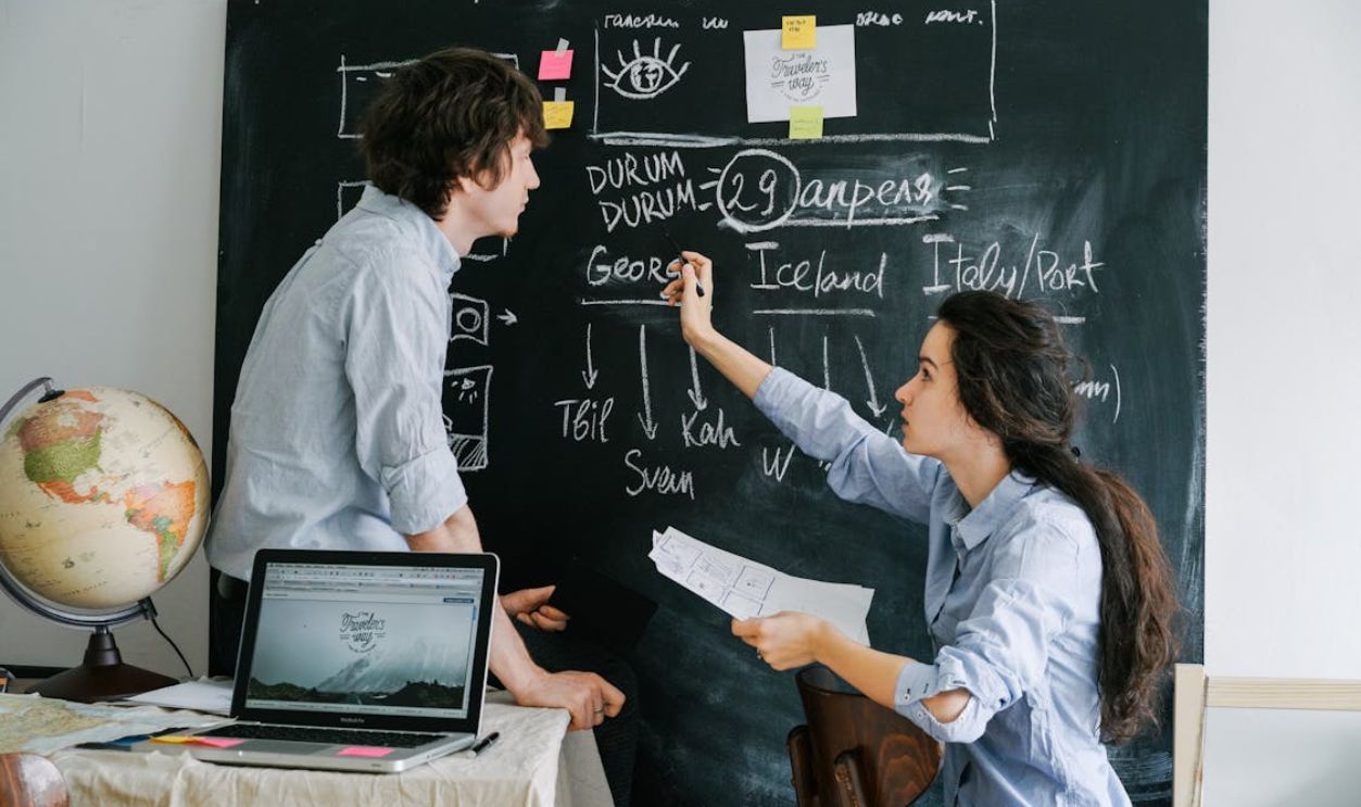 Two adults planning a trip using chalkboard and laptop in a casual indoor setting.