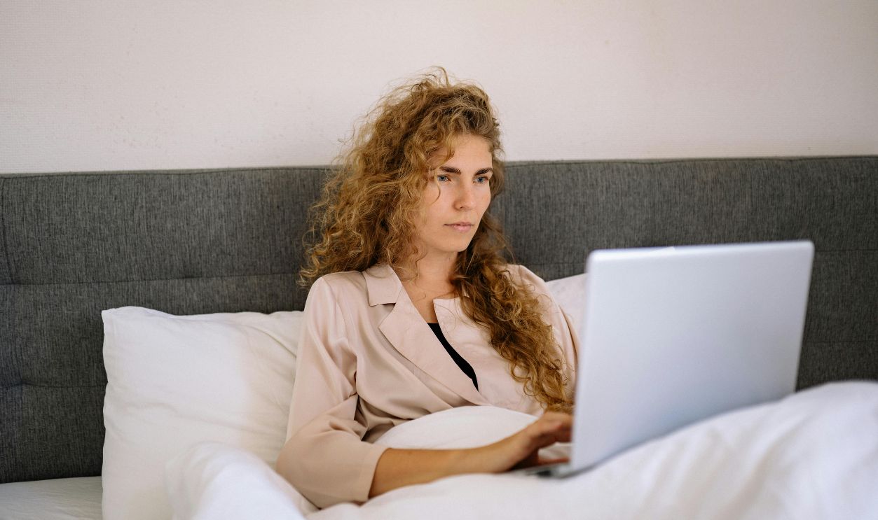 Focused woman with curly hair working on a laptop while sitting in bed in a cozy home setting.