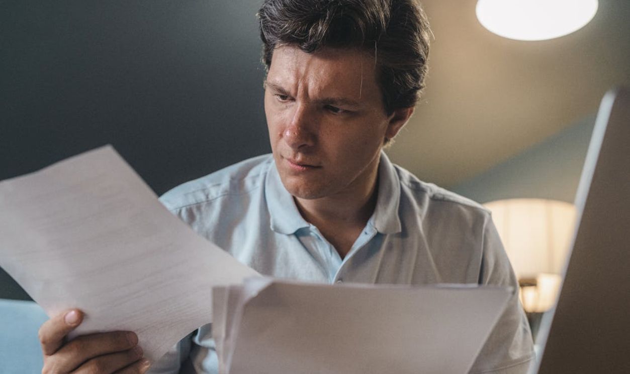 A man intently examines papers, seated indoors under warm lighting, focusing on his work.
