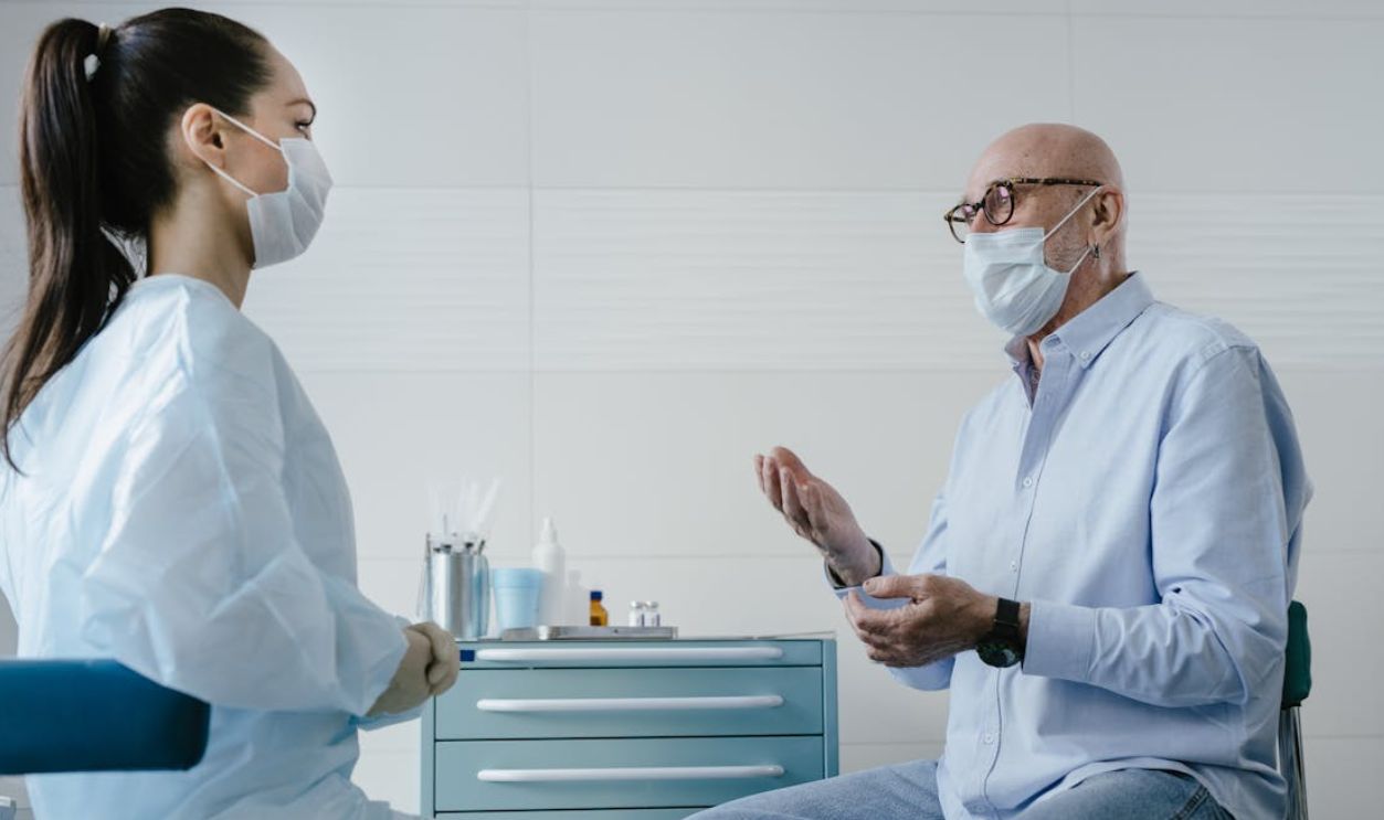 A senior adult patient discusses health concerns with a masked medical professional in a clinic setting.