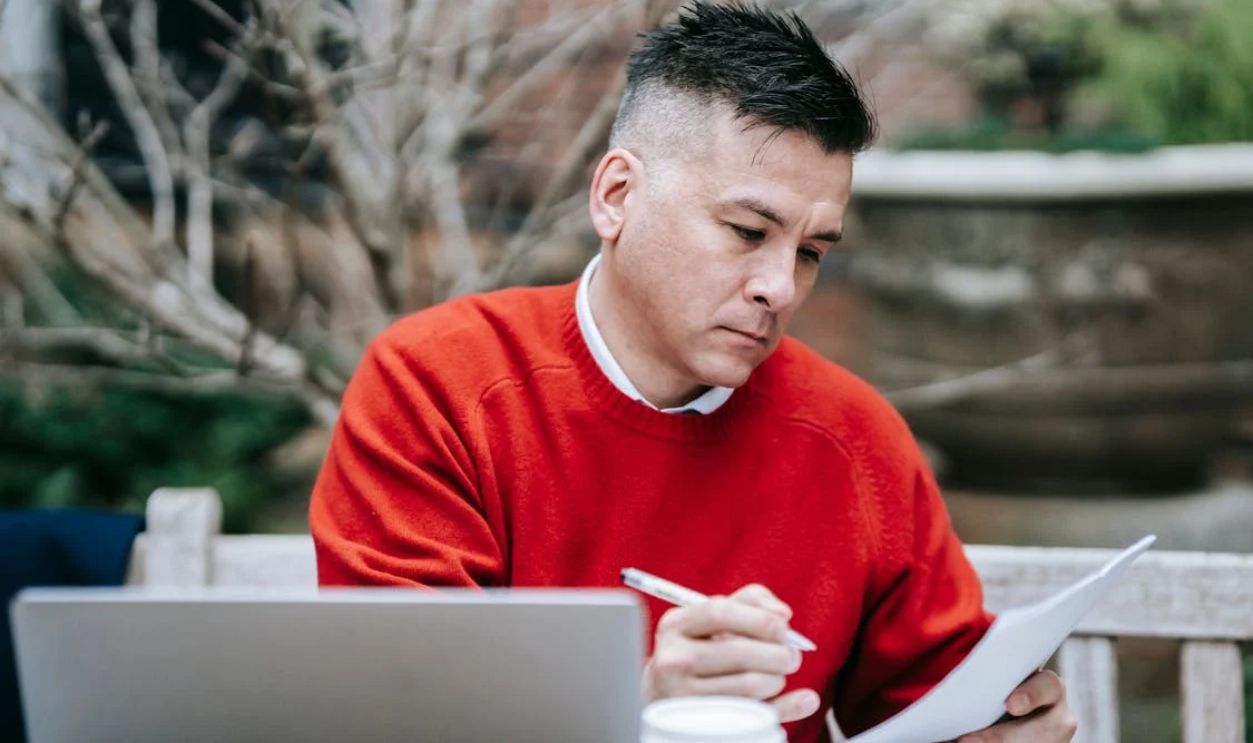 A man in a red sweater concentrating on paperwork while working remotely with a laptop outdoors.