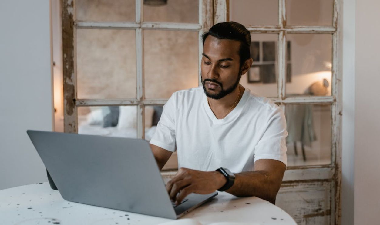 Adult man with beard focused on laptop work at home desk during the day.