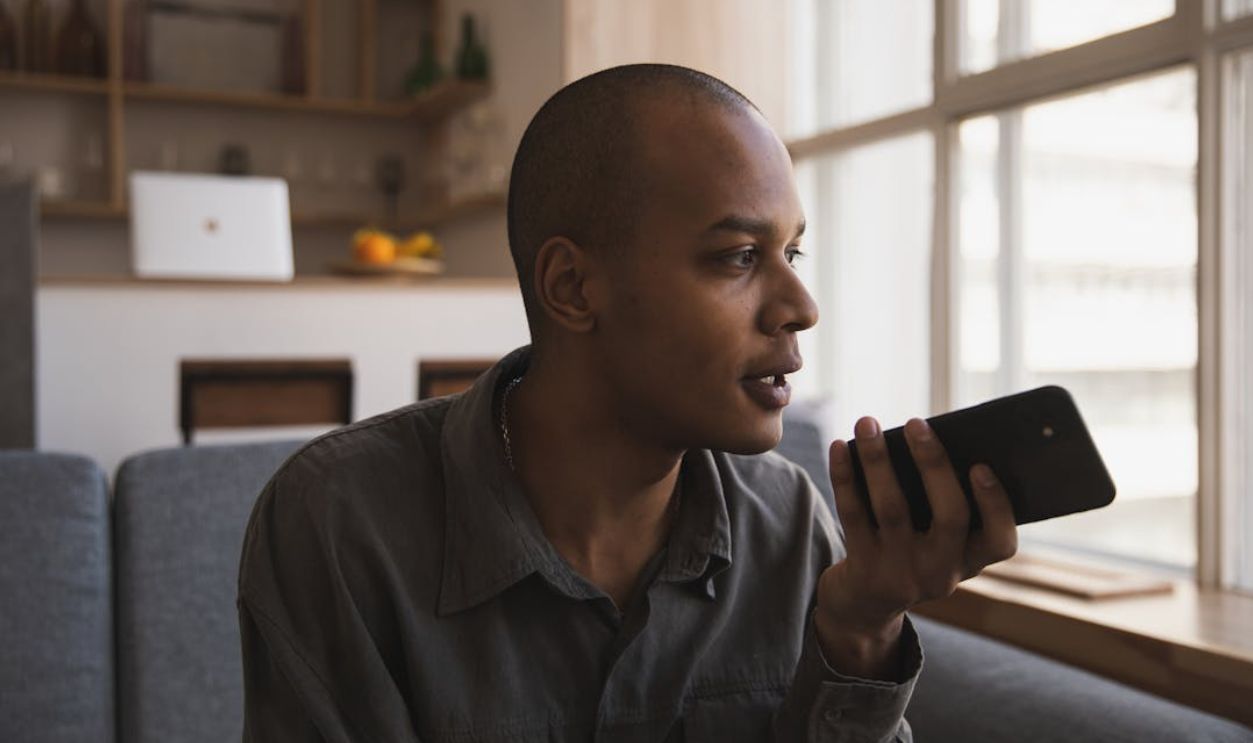 Man holding smartphone and speaking indoors, sitting on sofa by window lighting.