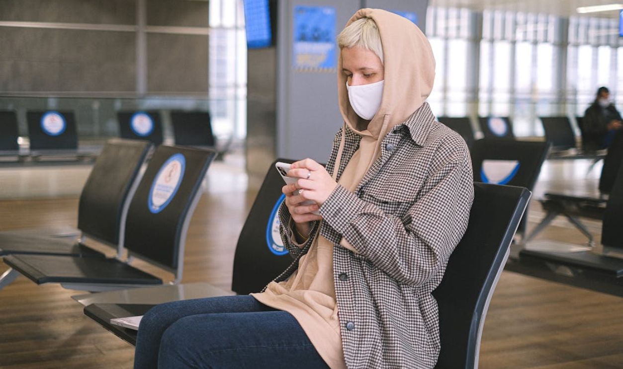 A woman wearing a face mask and a hood sits in an airport waiting area, using her smartphone.