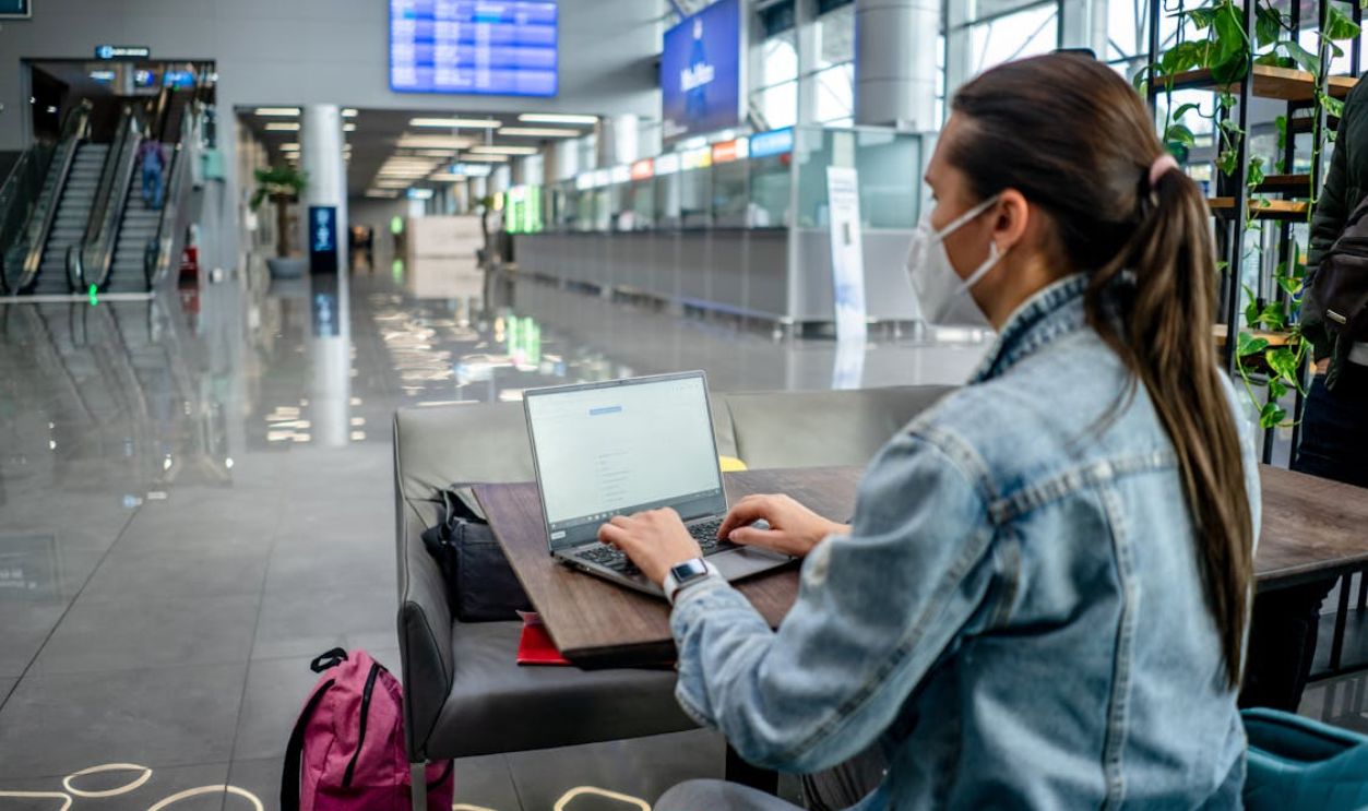 A woman wearing a mask works on her laptop inside a spacious airport terminal.