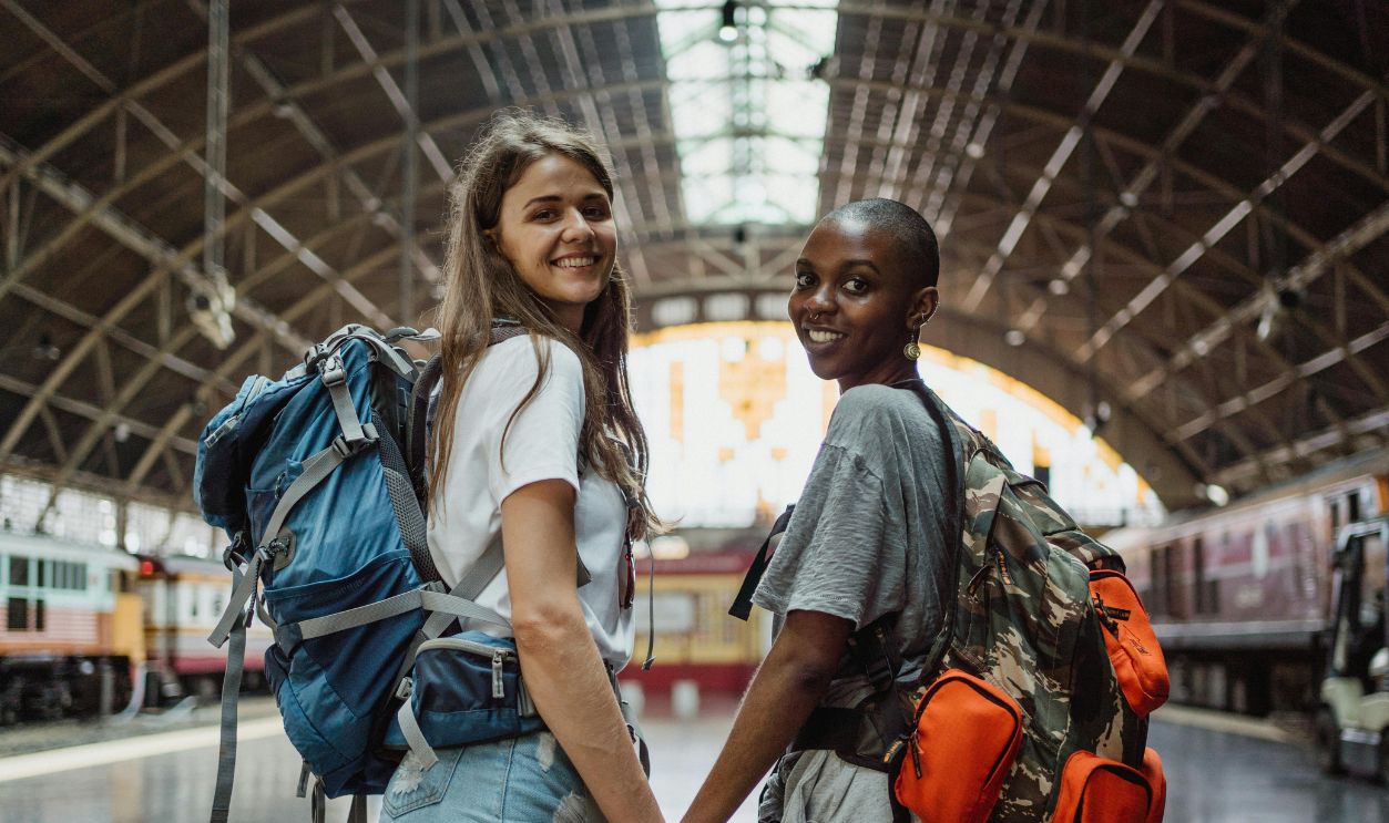 Two young women with backpacks holding hands, smiling at a railway station