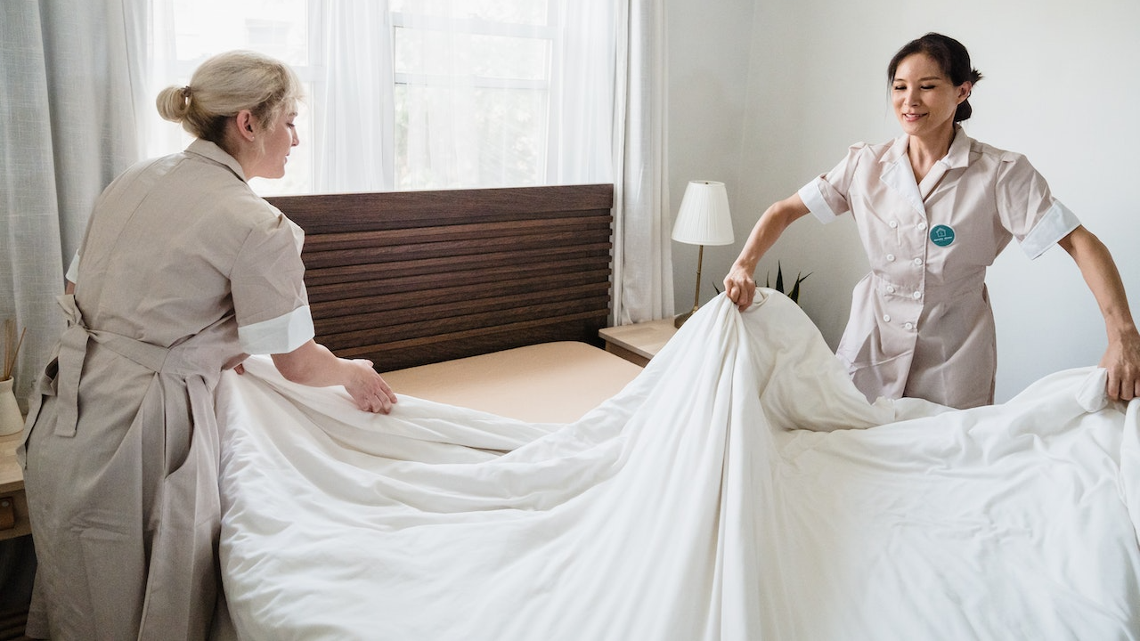 Housekeepers wearing uniforms changing bed sheets in hotel room.