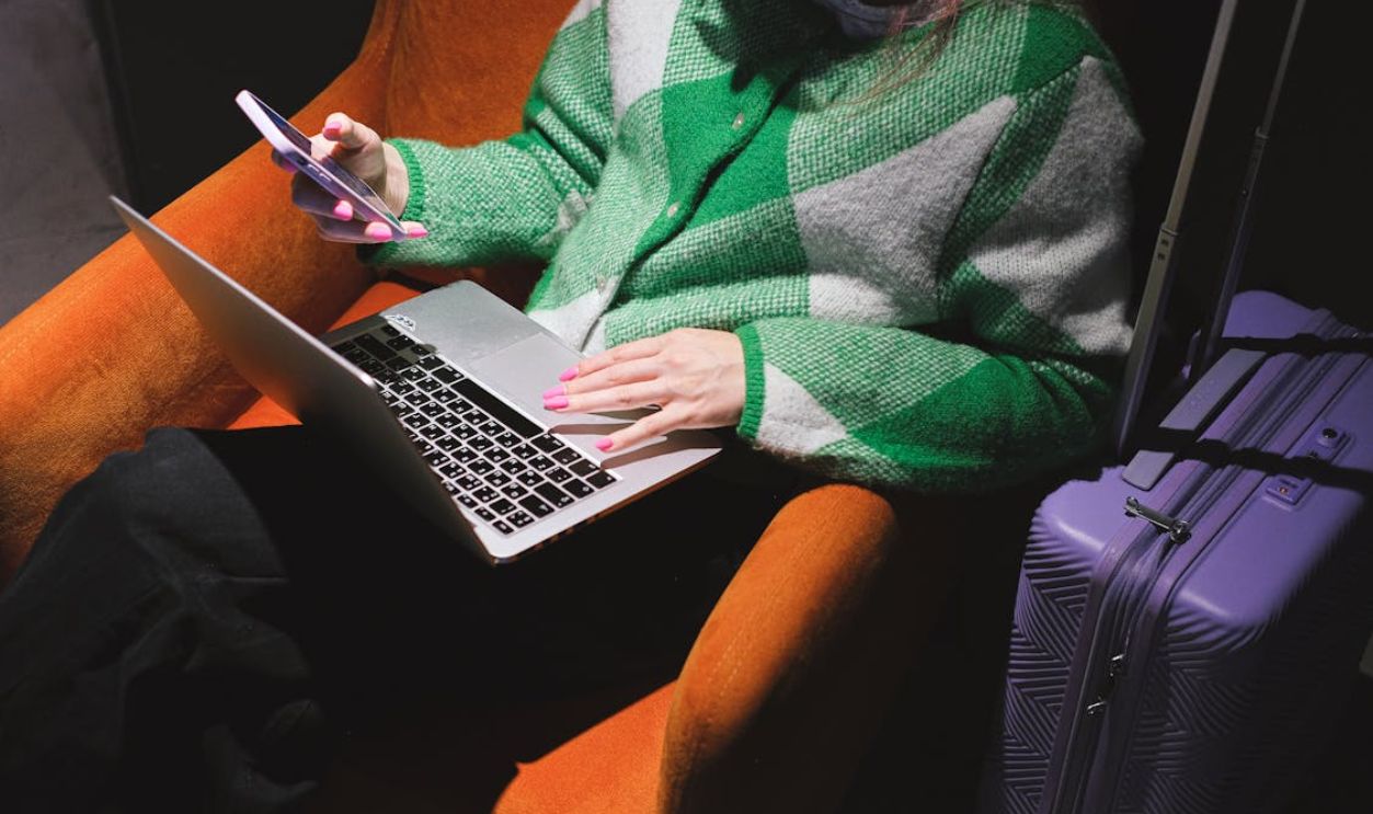 Woman in a knitted sweater working remotely on laptop with a smartphone, sitting in an armchair