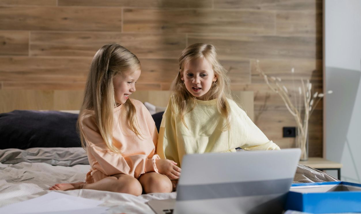 Two young girls learning online together at home, sitting on a bed 