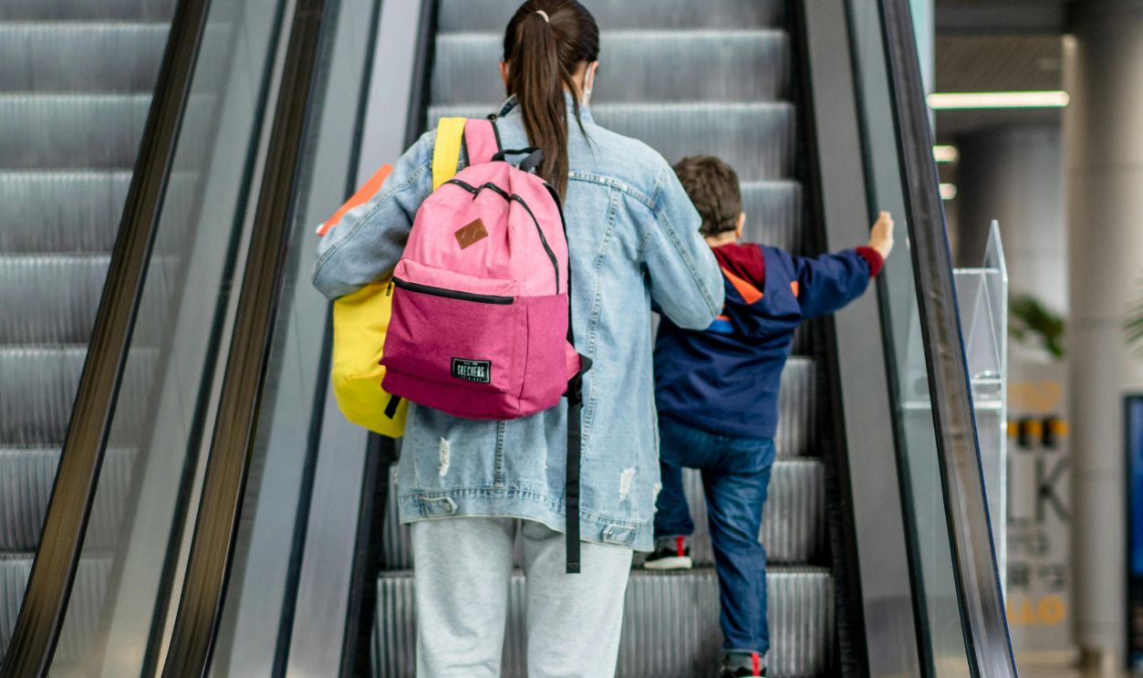 A mother with a backpack and her child ascend an escalator inside a shopping mall.