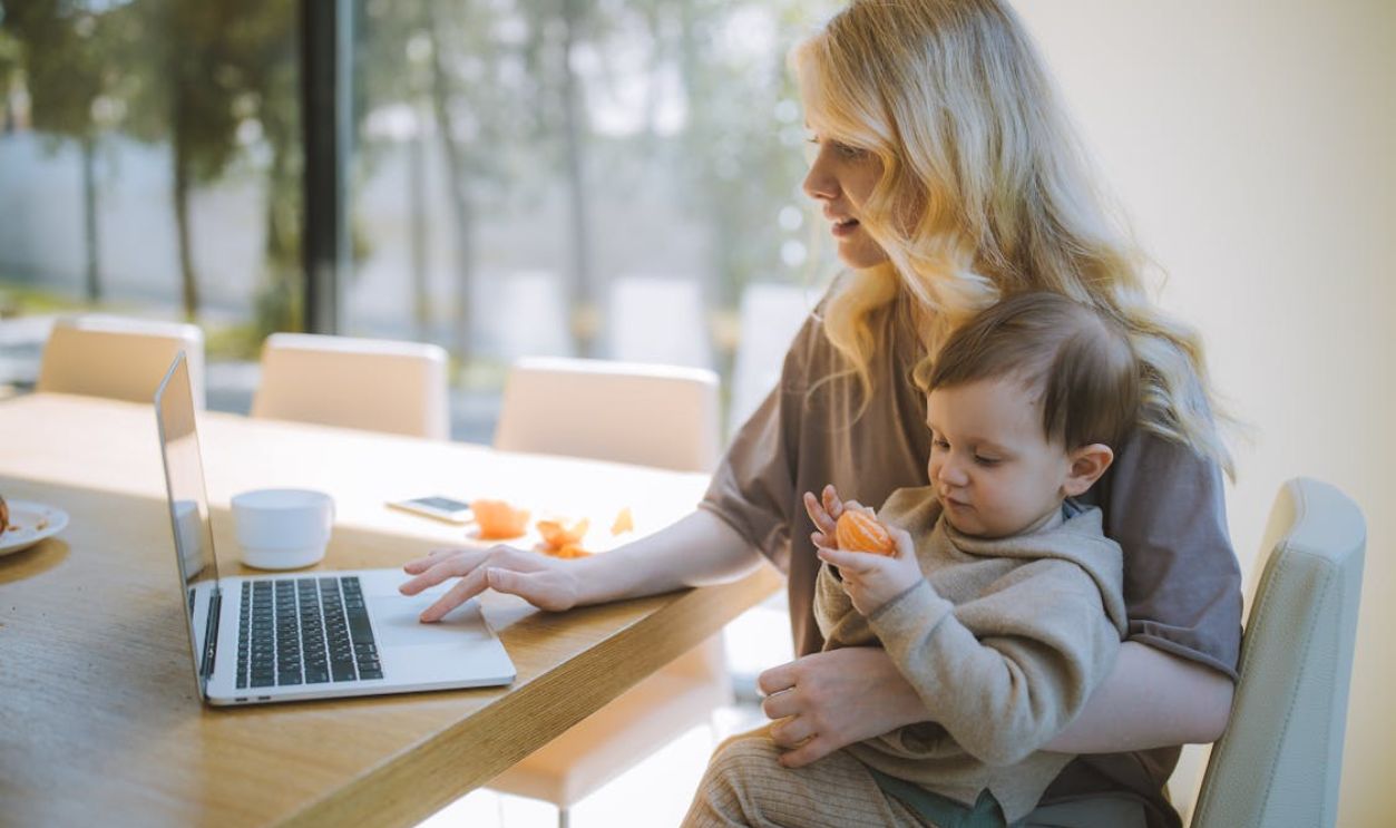A mother works on a laptop at home while holding her baby, showcasing remote working and parenting