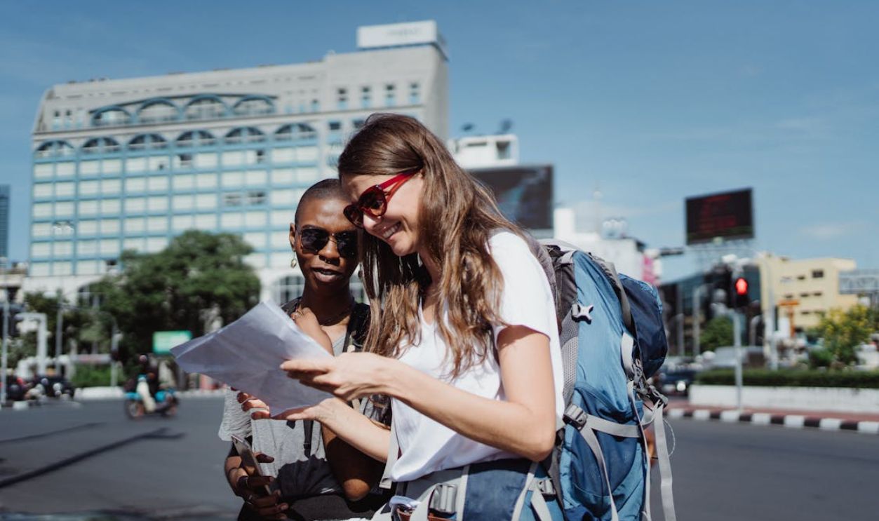 Two women tourists with backpacks check a map on a city street under a bright sky.