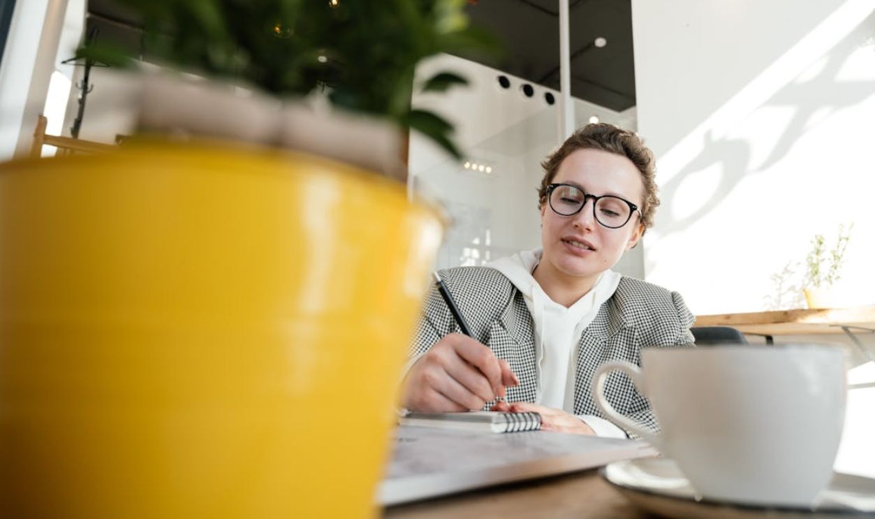 A young woman in a cafe jotting notes, framed by a potted plant and coffee cup.