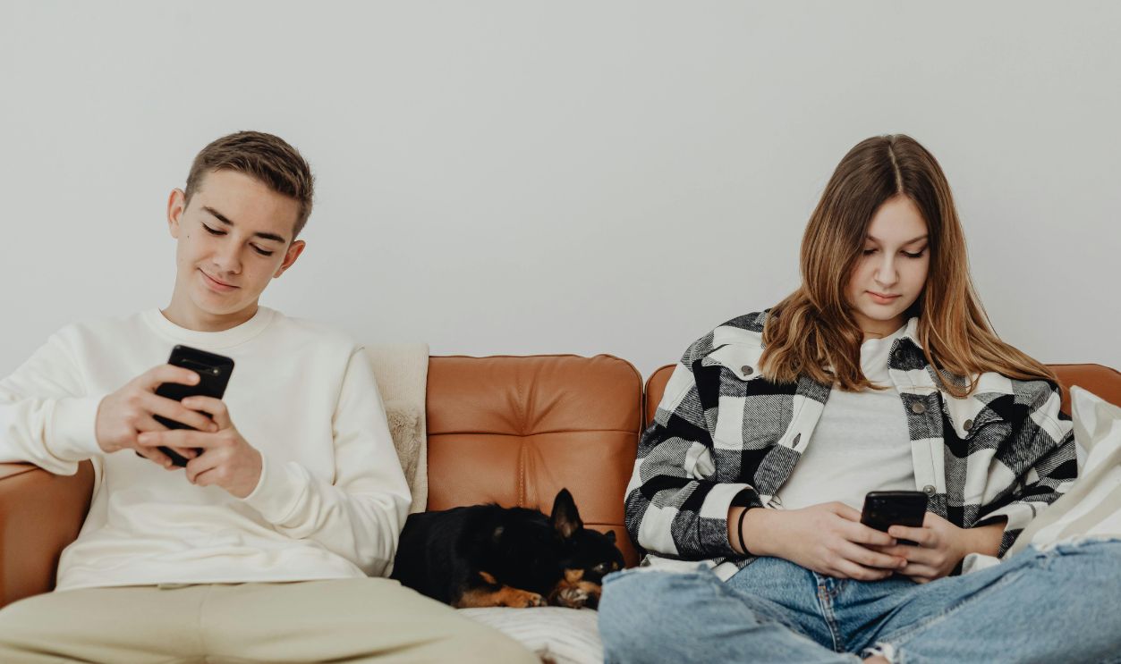 Two teenagers sitting on a sofa with a dog, using smartphones 