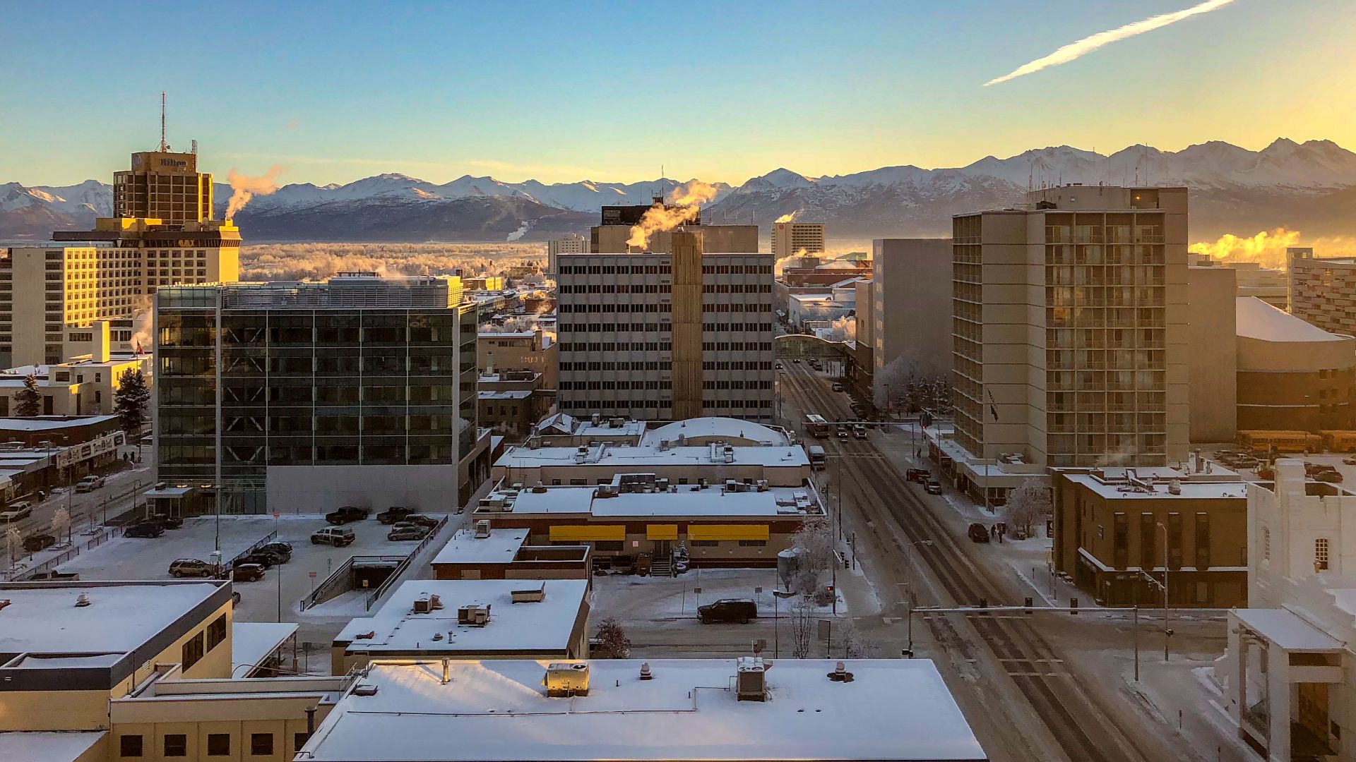 The sun sets on Downtown Anchorage on a cold, quiet day in February, with the Chugach Mountain Range just below the horizon above snow-covered rooftops, as seen from the Hotel Captain Cook.