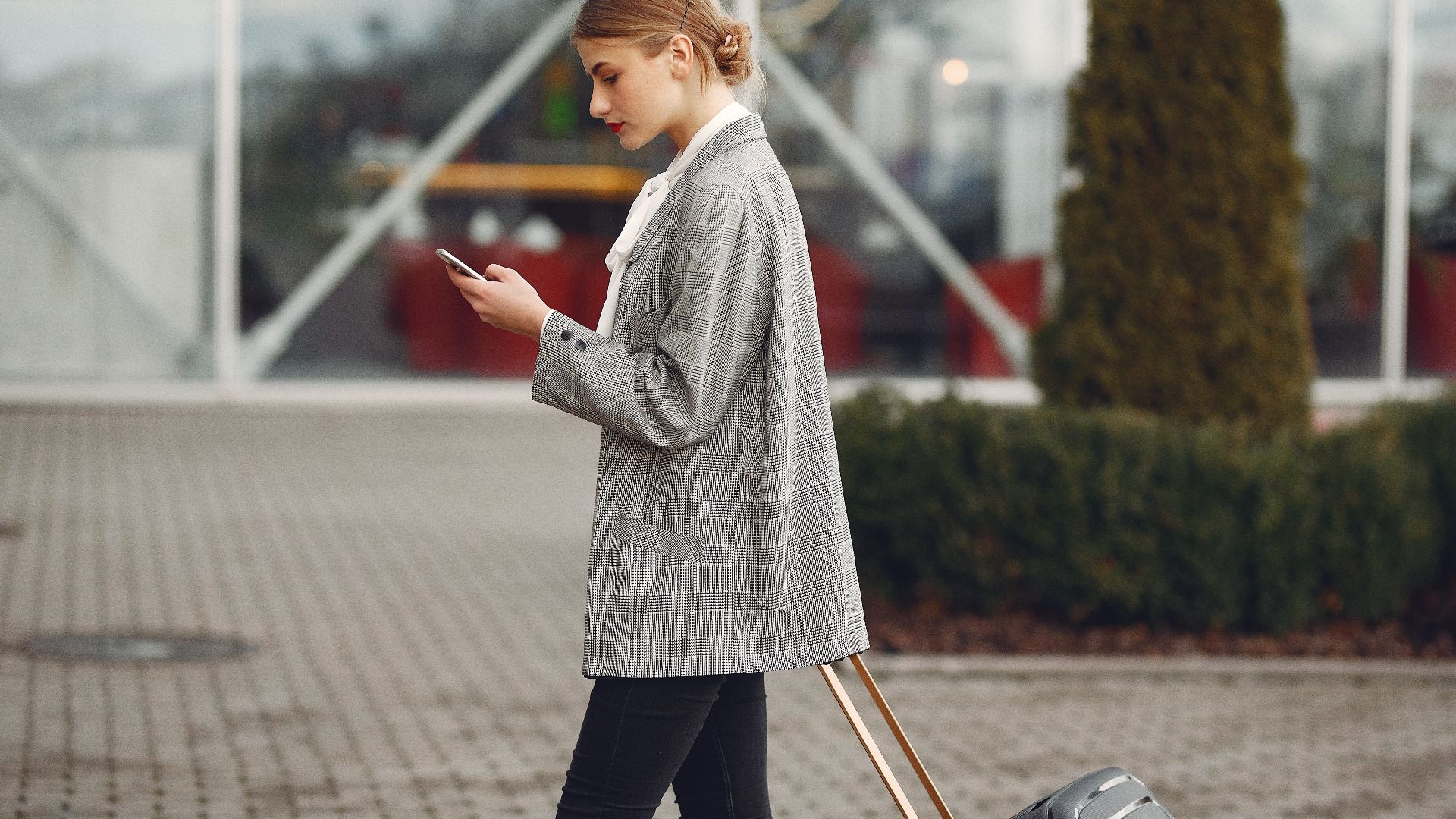 Stylish woman walking with suitcase while using smartphone outside a modern building.