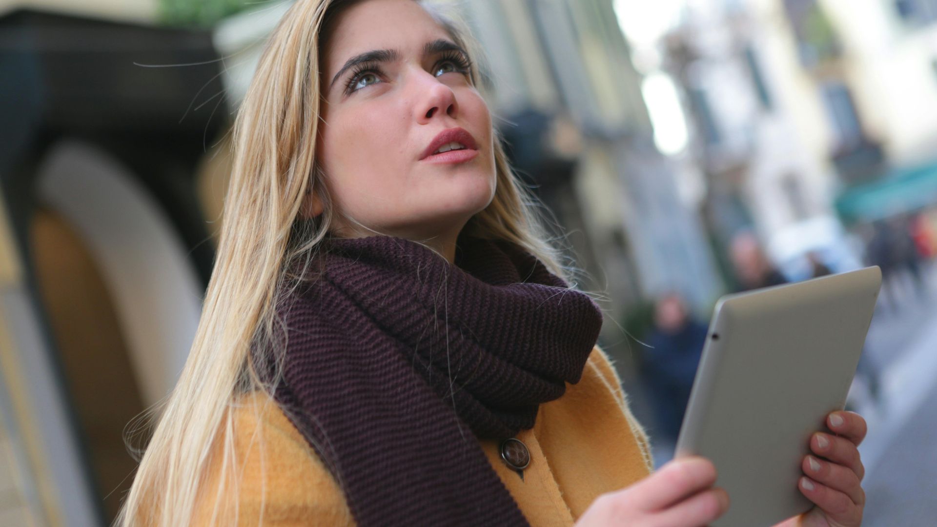 A young woman with a tablet looks up thoughtfully in a vibrant city street.