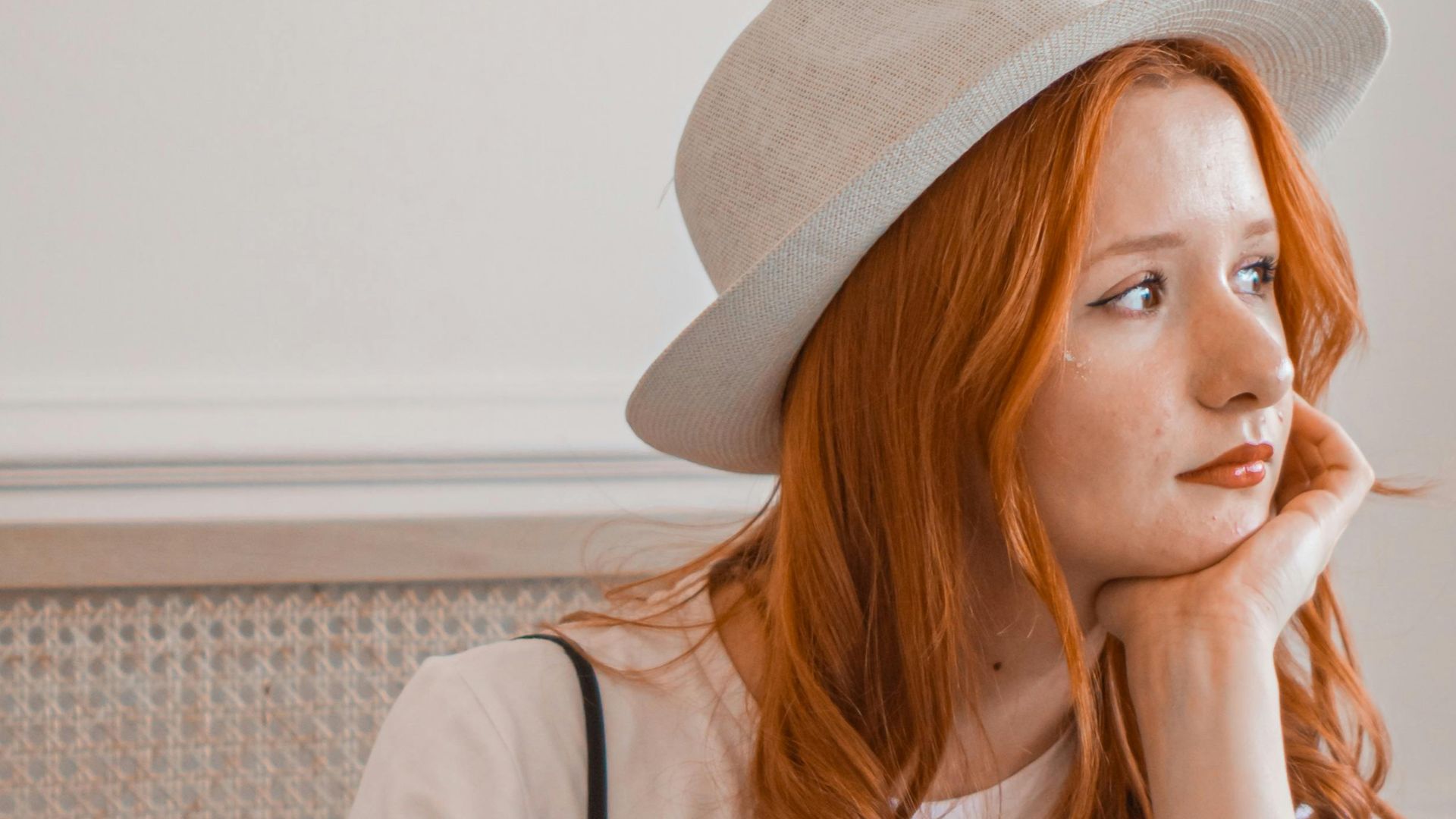 Redhead woman in a straw fedora using a laptop indoors, deep in thought.