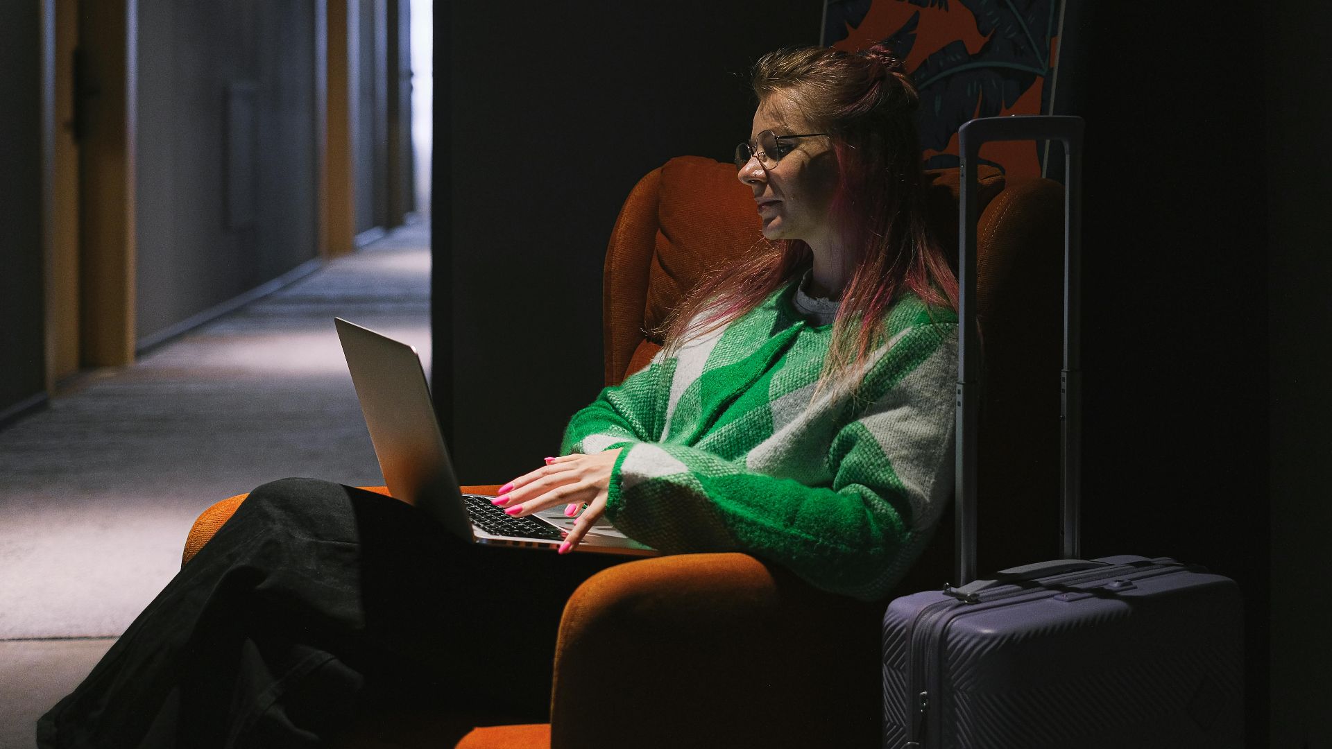 A woman in a hotel hallway using a laptop, sitting on an armchair next to luggage, creating a cozy working environment.
