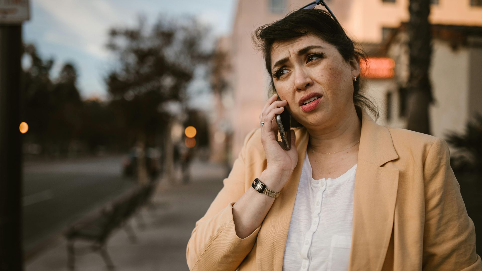 Woman in beige jacket looks concerned while making phone call outdoors.