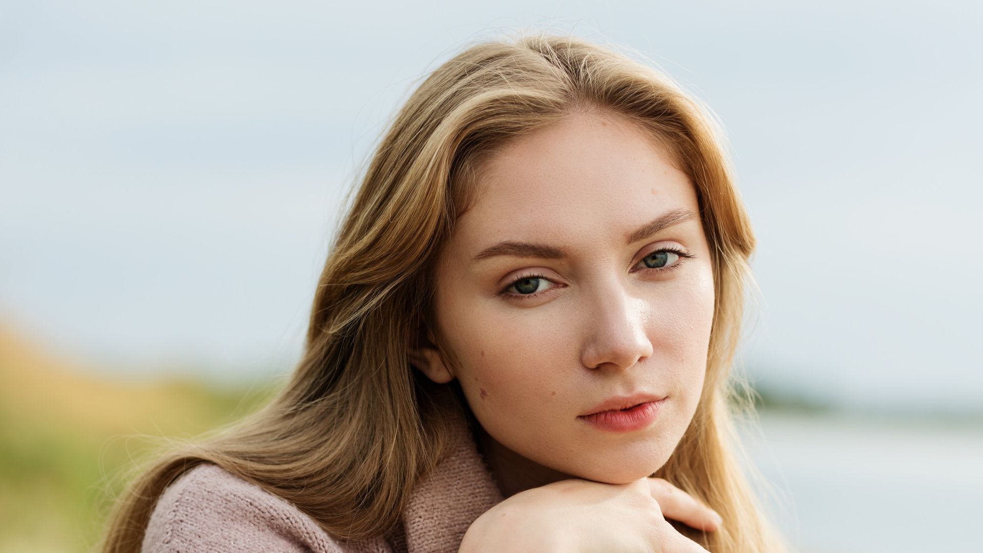 Caucasian woman sits by the beach in a sweater, lost in thought on a breezy day.