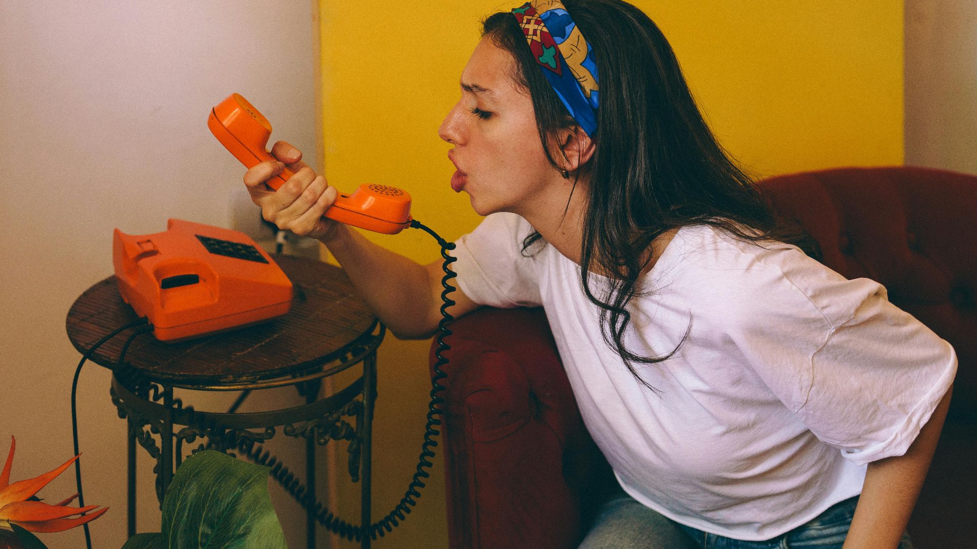 Woman speaking into a bright orange rotary phone indoors, expressing emotion.