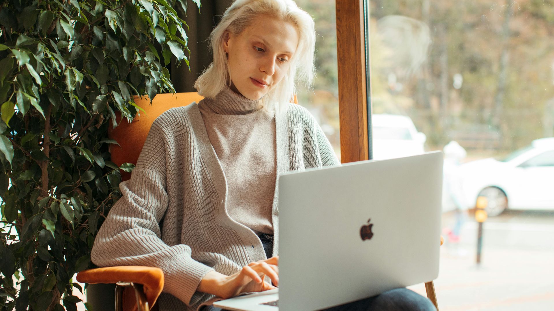 Blonde woman working on a laptop in a cozy office setting with indoor plants.