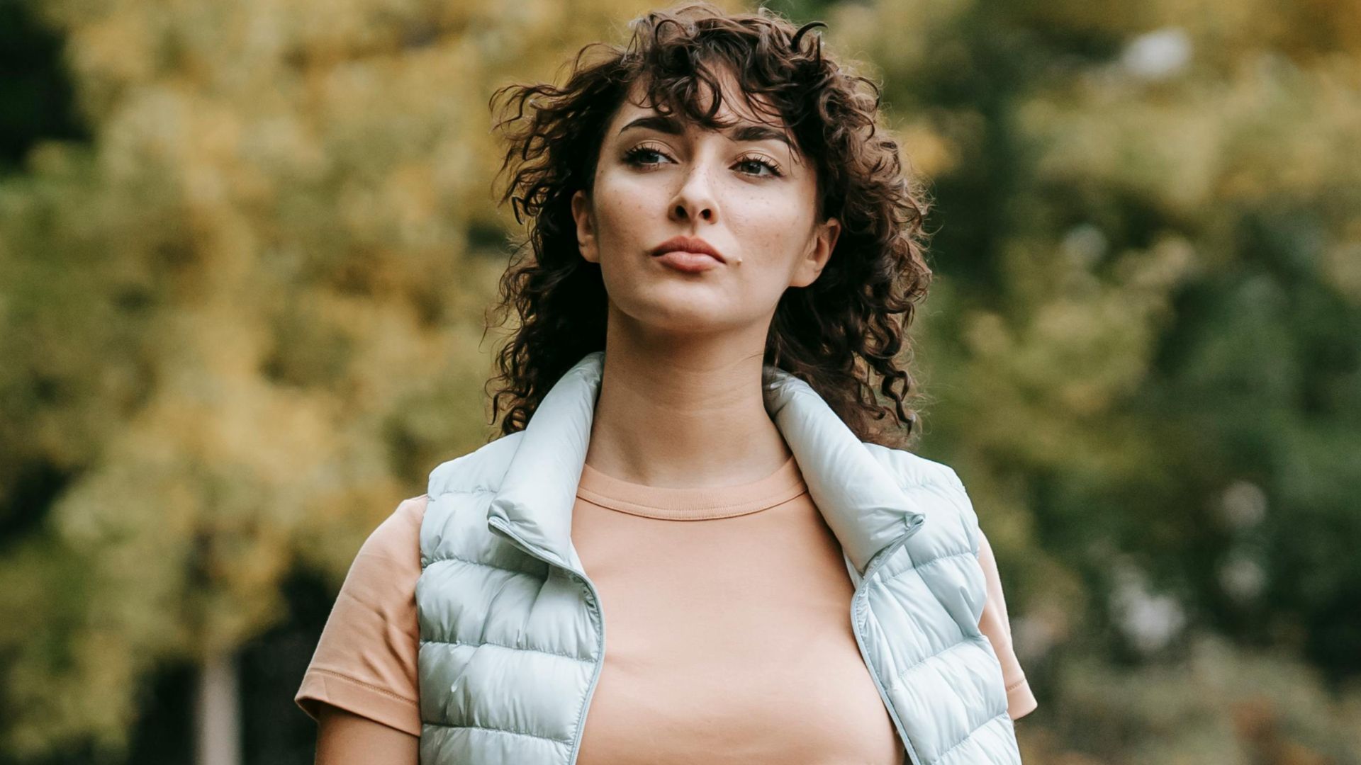 Pensive woman in casual clothes walking alone on walkway near green trees placed in city park and looking away in daytime