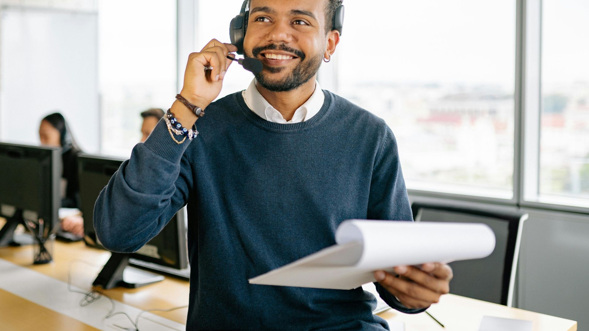 A cheerful man wearing a headset and holding a clipboard at a modern office workspace.