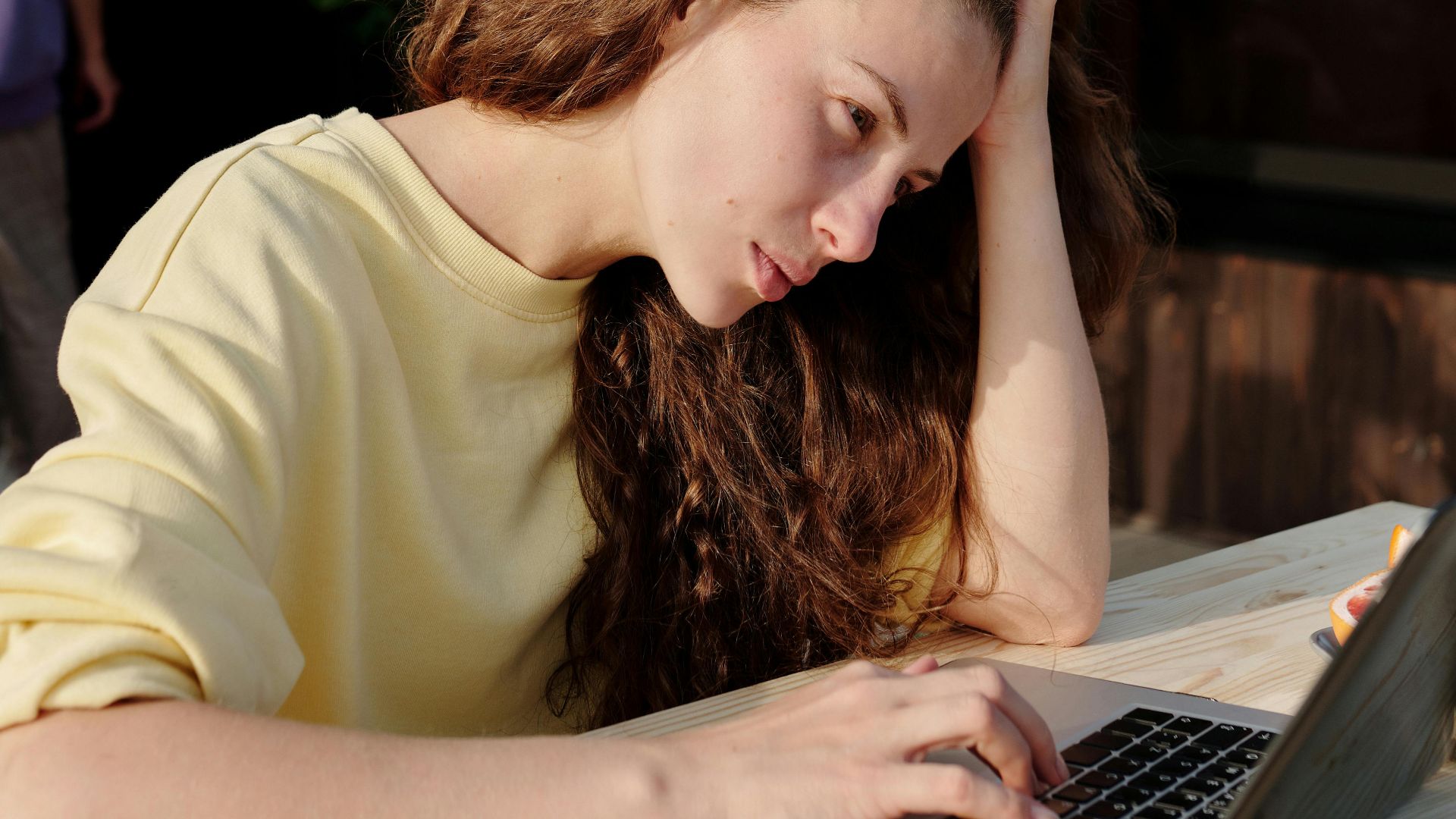A young woman with long hair works outdoors on a laptop, appearing focused and thoughtful.