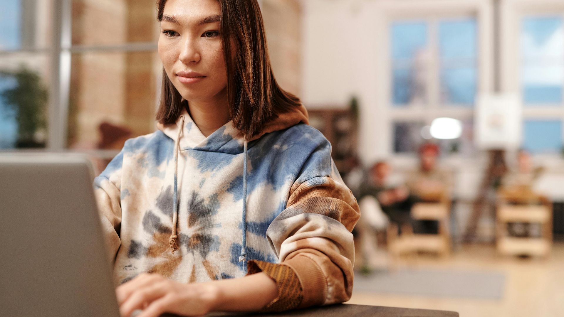 Asian woman in tie-dye hoodie working on laptop at wooden table in bright modern office.
