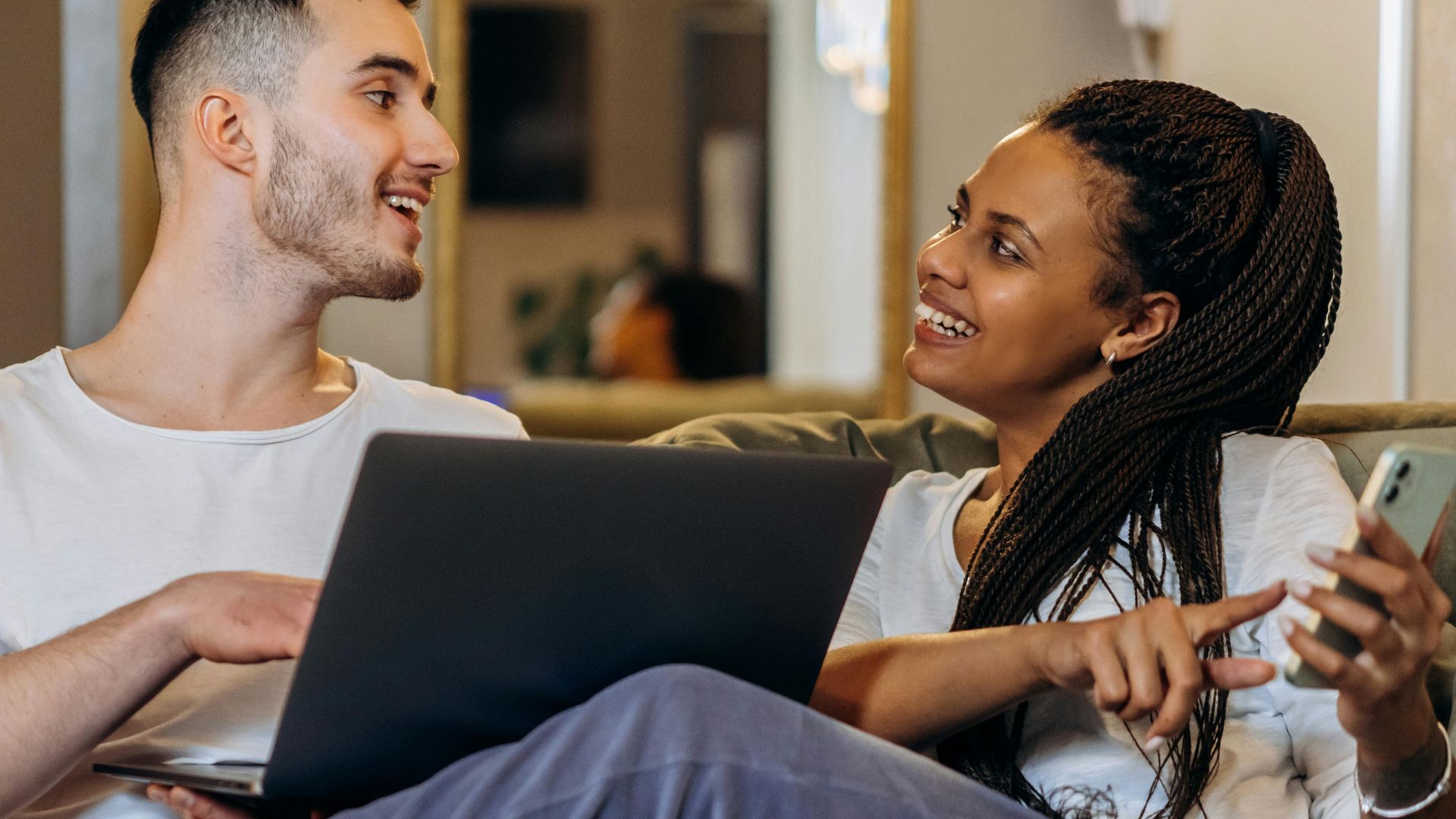 Interracial couple bonding over technology, sitting on a couch indoors with a laptop and smartphone in hand.