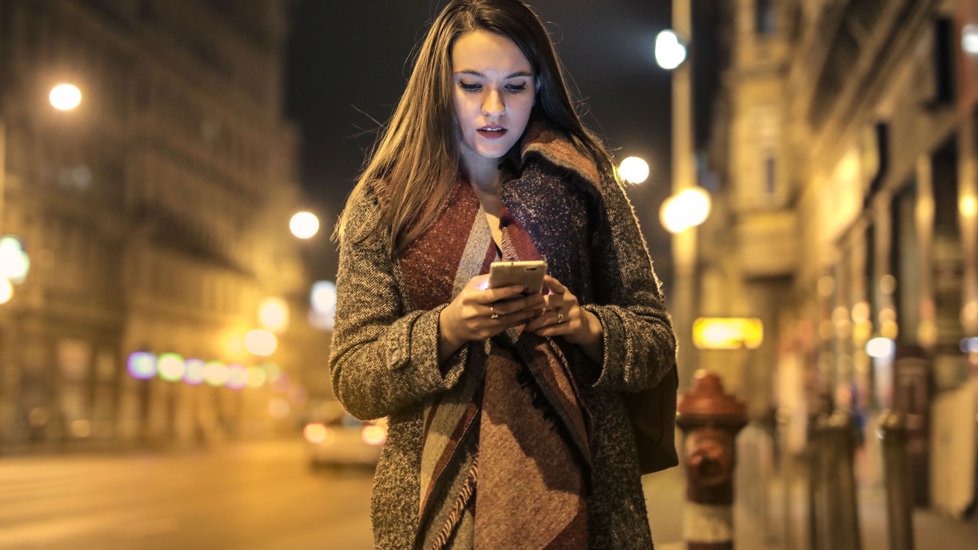 Elegant woman walks through urban street at night, engaged with her smartphone, surrounded by city lights.