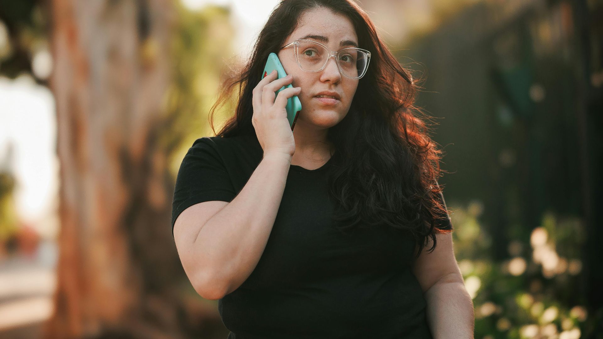 Woman with eyeglasses talks on smartphone in sunny park setting.