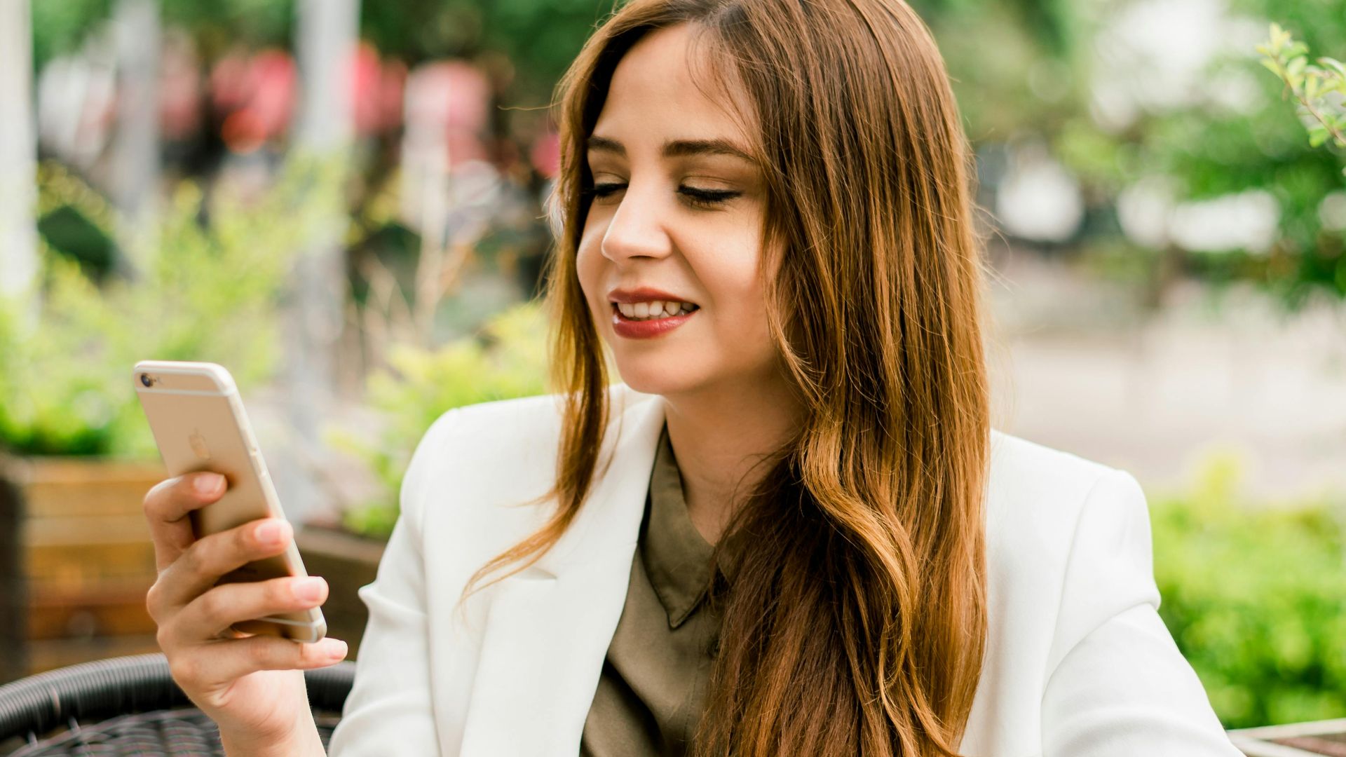 Smiling woman in a blazer uses smartphone while enjoying coffee outdoors in Guadalajara.