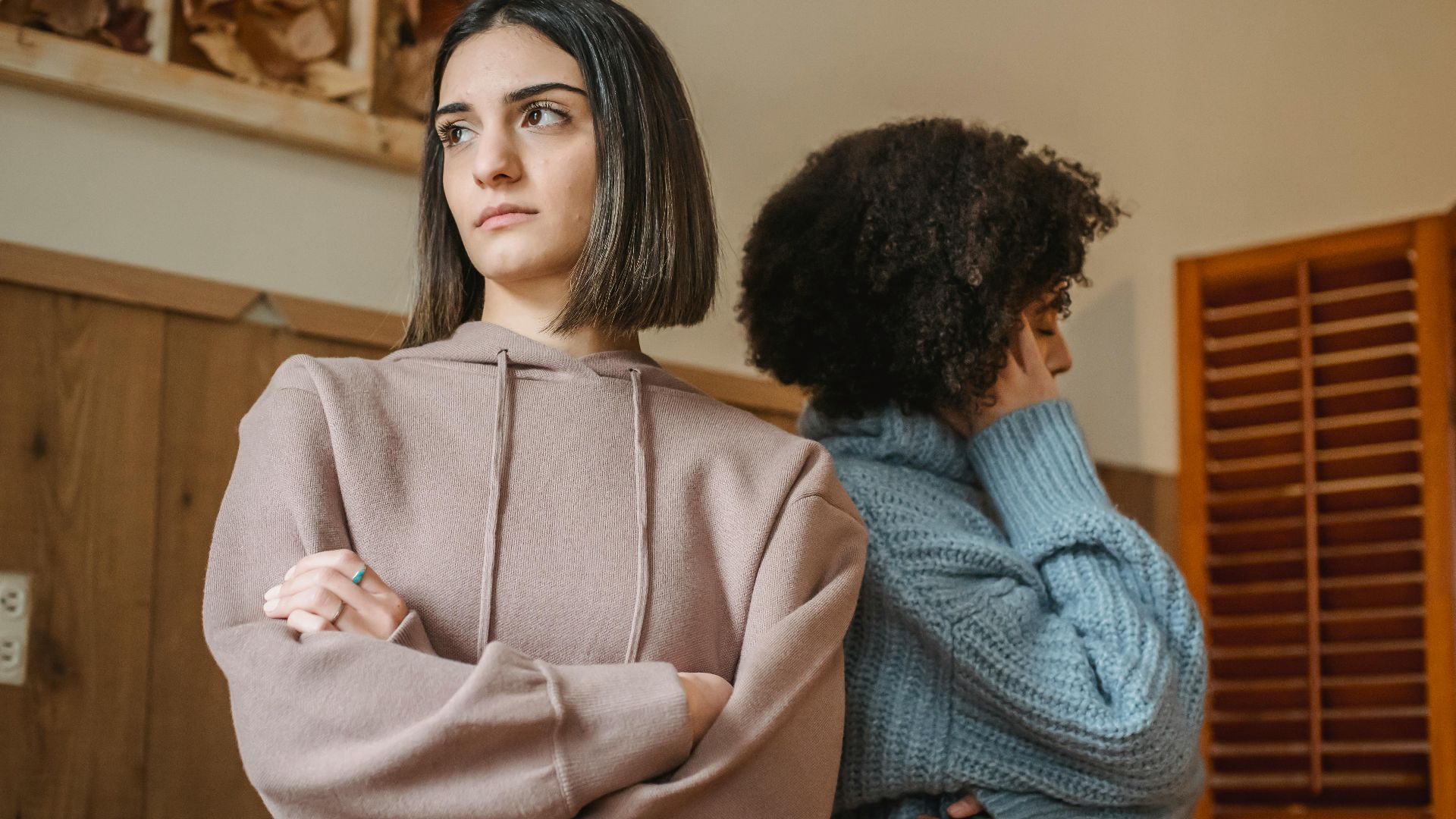 From below of unsatisfied multiracial women in casual clothes with crossed arms standing in light room during quarrel at home