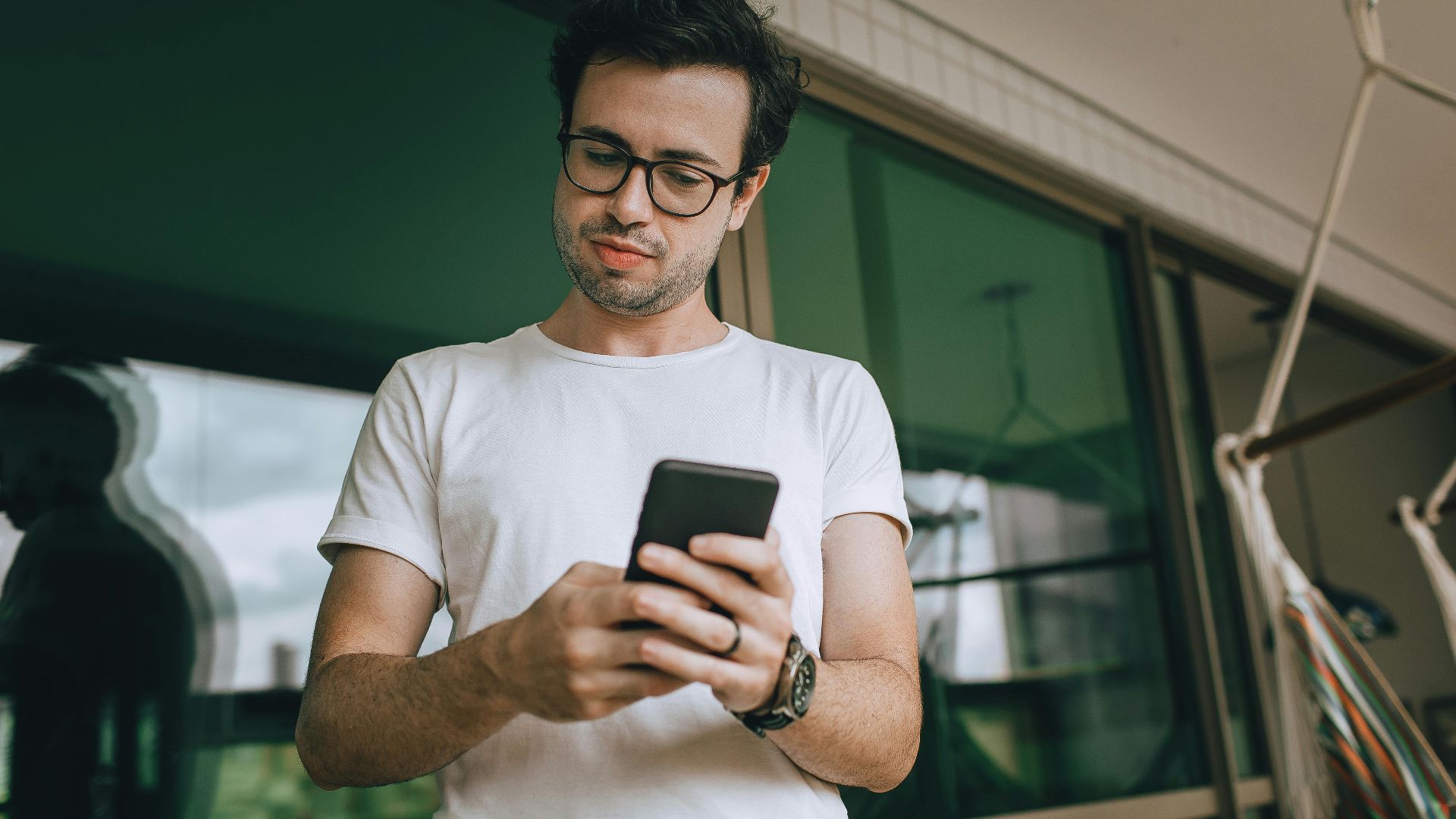 A young man in glasses texts on a smartphone outdoors on a modern balcony.