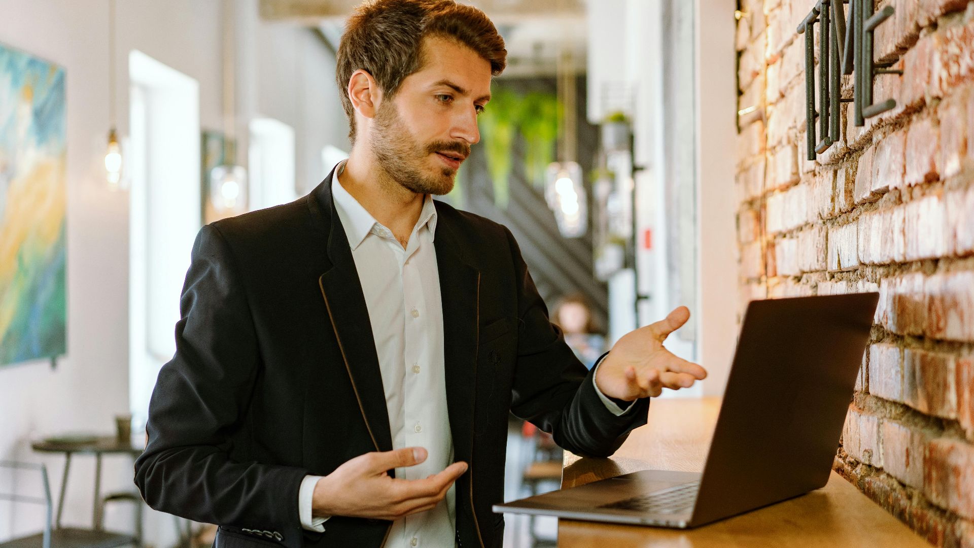 Young man in a black jacket engaged in an online meeting at a trendy cafe, gesturing while looking at the laptop.