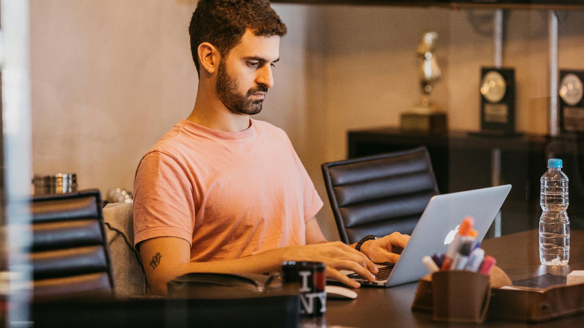 Man with short hair in pink t-shirt working on a laptop in a modern office setting.