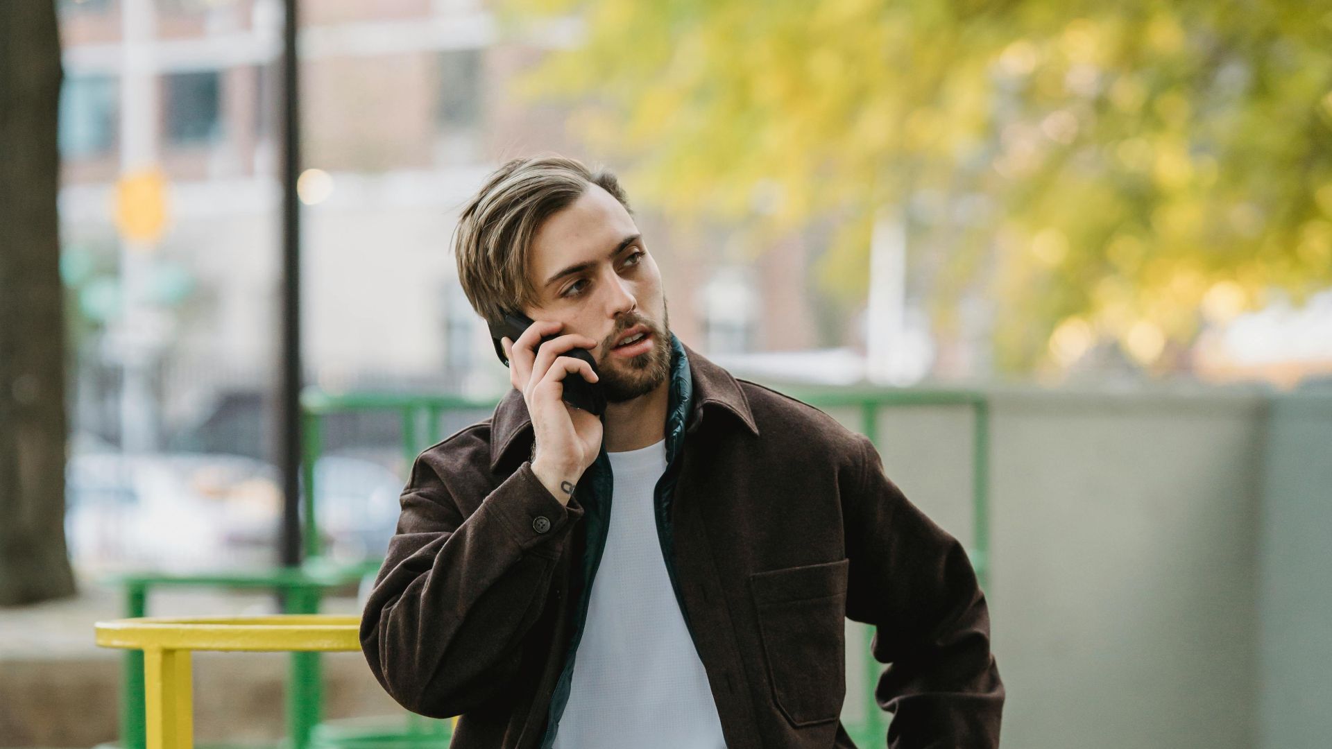 Young contemplative male in casual apparel with hand on hip speaking on cellphone while looking away in town