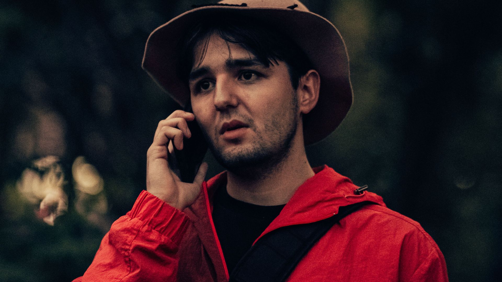 A man in a red jacket and hat talking on a phone while holding a camera in a forest setting.